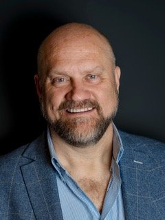 A middle-aged man with a bald head and beard, smiling, wearing a blue blazer and shirt, against a dark background.
