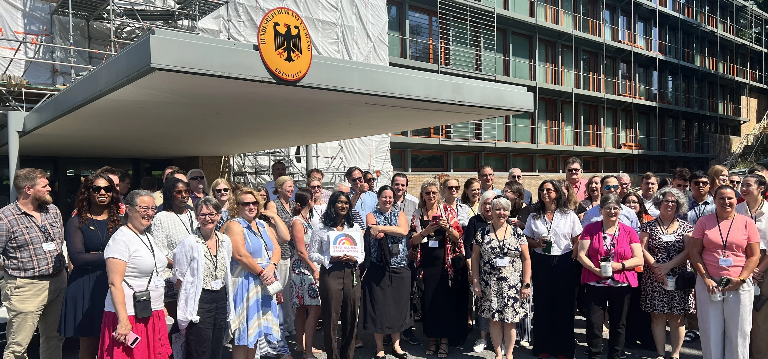 A large group of people gathered outdoors in front of a building with many windows, celebrating an alumni event, with a logo of the German embassy visible on a sign above.