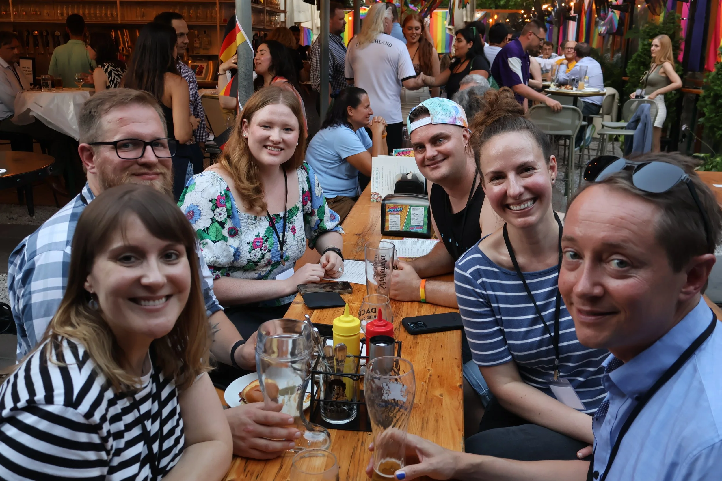 A group of people sitting at a wooden table in a lively outdoor party or festival, with colorful decorations and numerous other attendees in the background. The group is smiling and enjoying drinks, with some wearing casual summer clothing and sungla