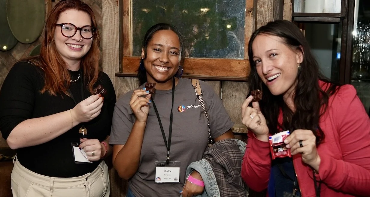 Three women smiling and holding chocolate bars indoors at a social gathering.