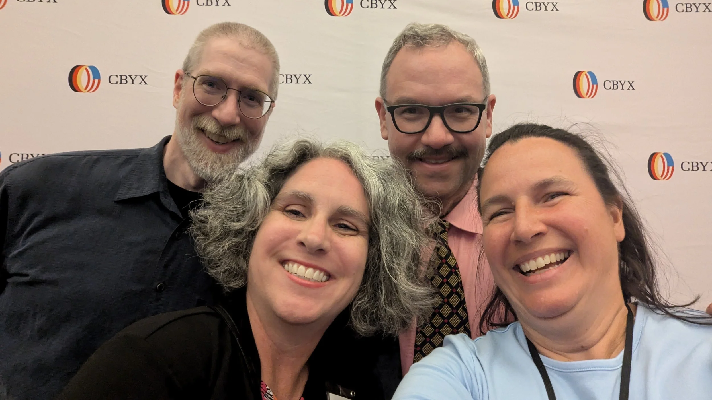 Group of five people smiling for a selfie in front of a white backdrop with a logo and text. The group includes two men with glasses and glasses, and three women, all smiling joyfully.