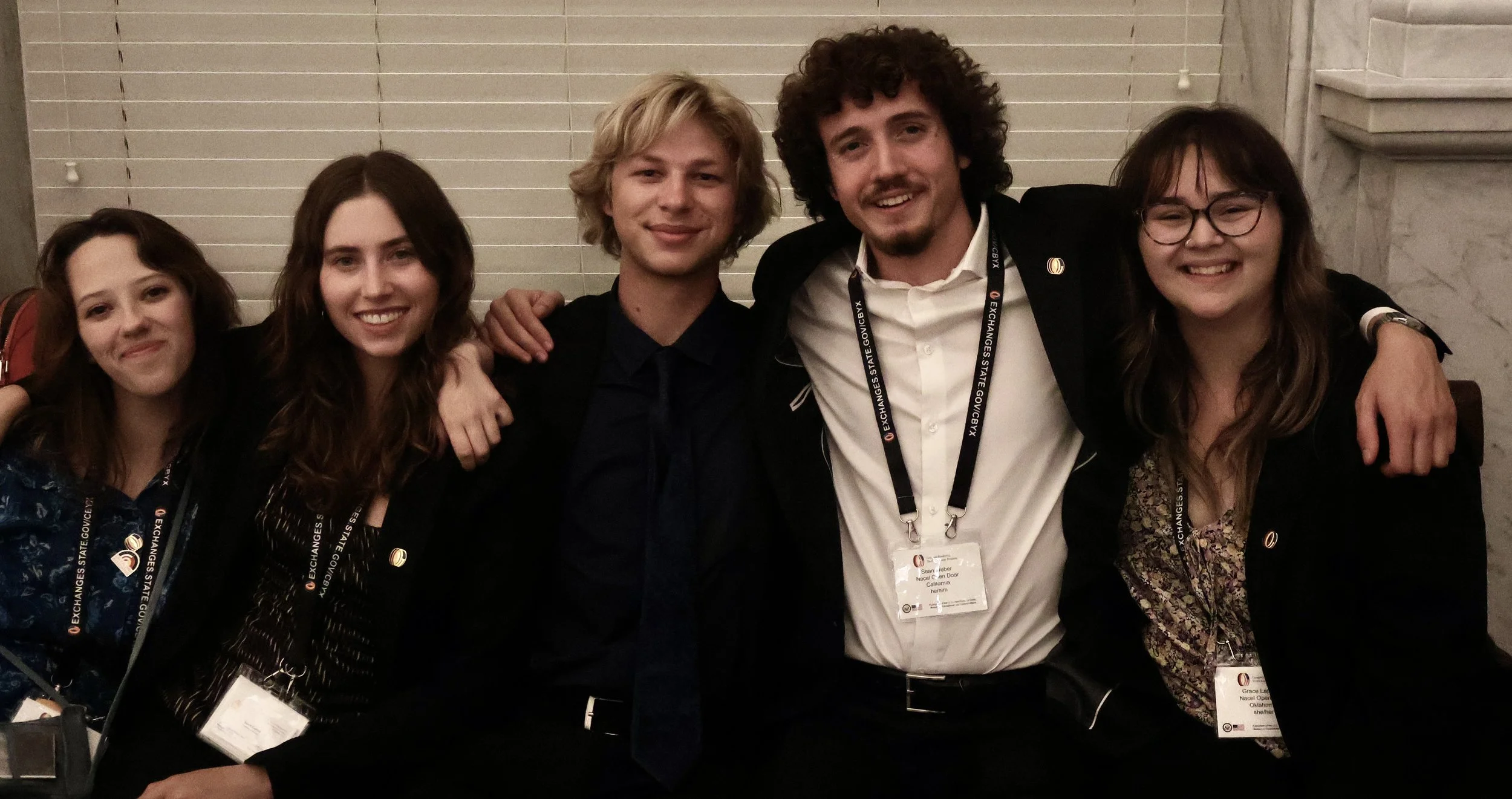 Five young adults smiling and posing together at a conference, with badges around their necks and their arms around each other, standing against a background of beige blinds and marble walls.