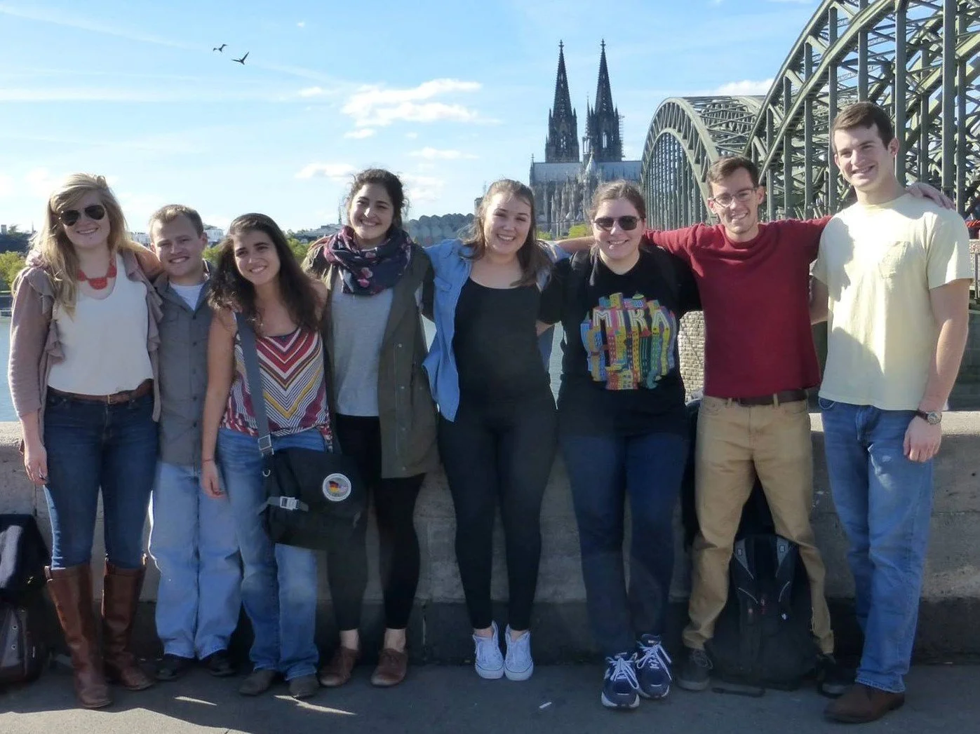 Group of eight people standing together outdoors with a bridge and a cathedral in the background, smiling and posing for a photo.