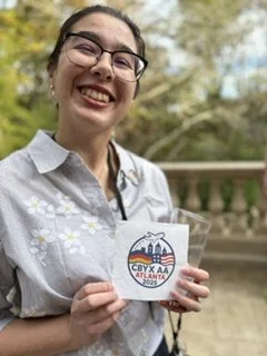 Young woman smiling outdoors, holding a sign with a cartoon pig and the logo of CBYX AA program.