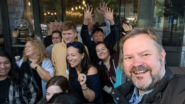 Group of smiling people taking a selfie outdoors, some waving, at a social gathering or event.