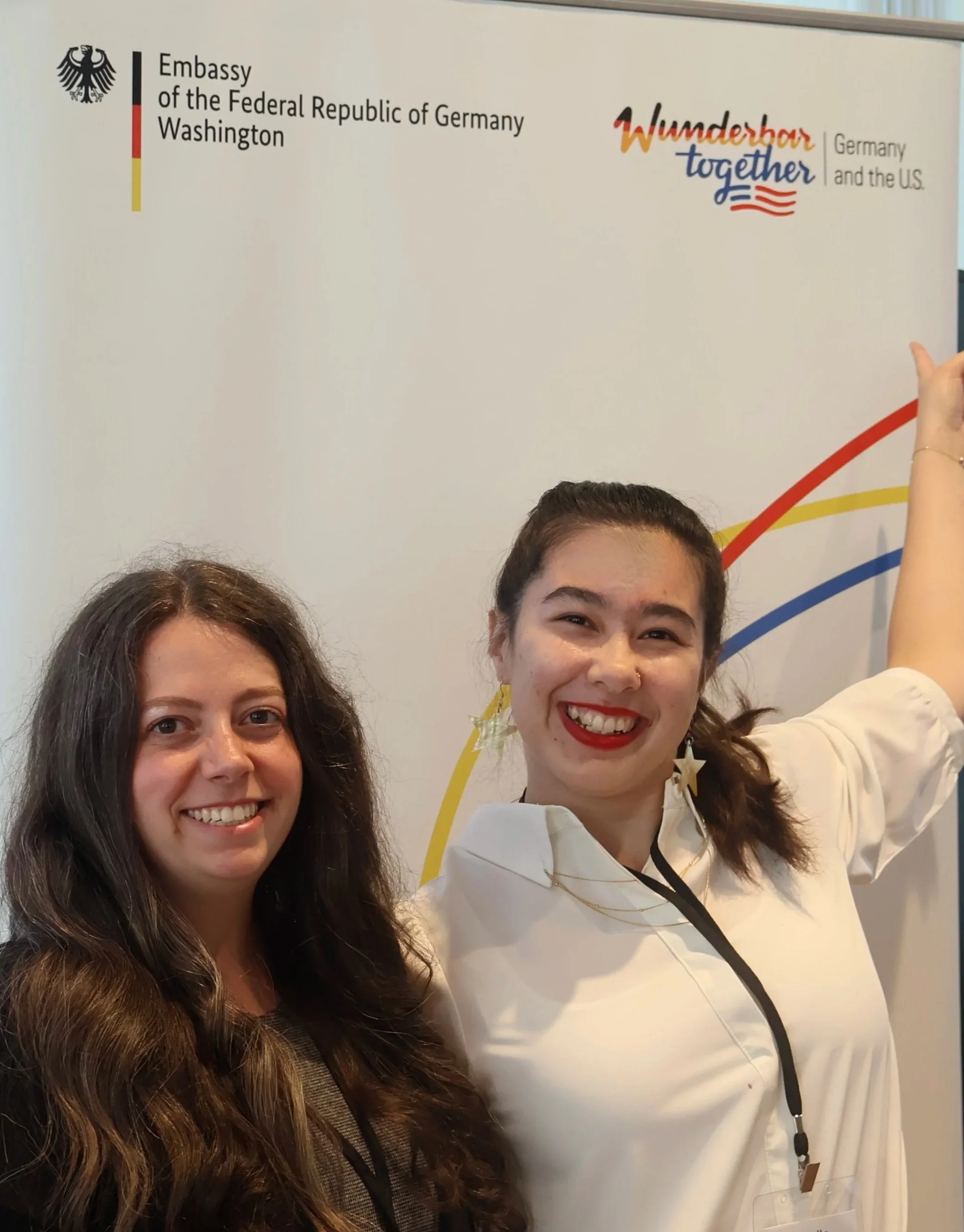 Two women smiling and taking a selfie in front of a sign that reads 'Embassy of the Federal Republic of Germany Washington' and displays the German and American flags.