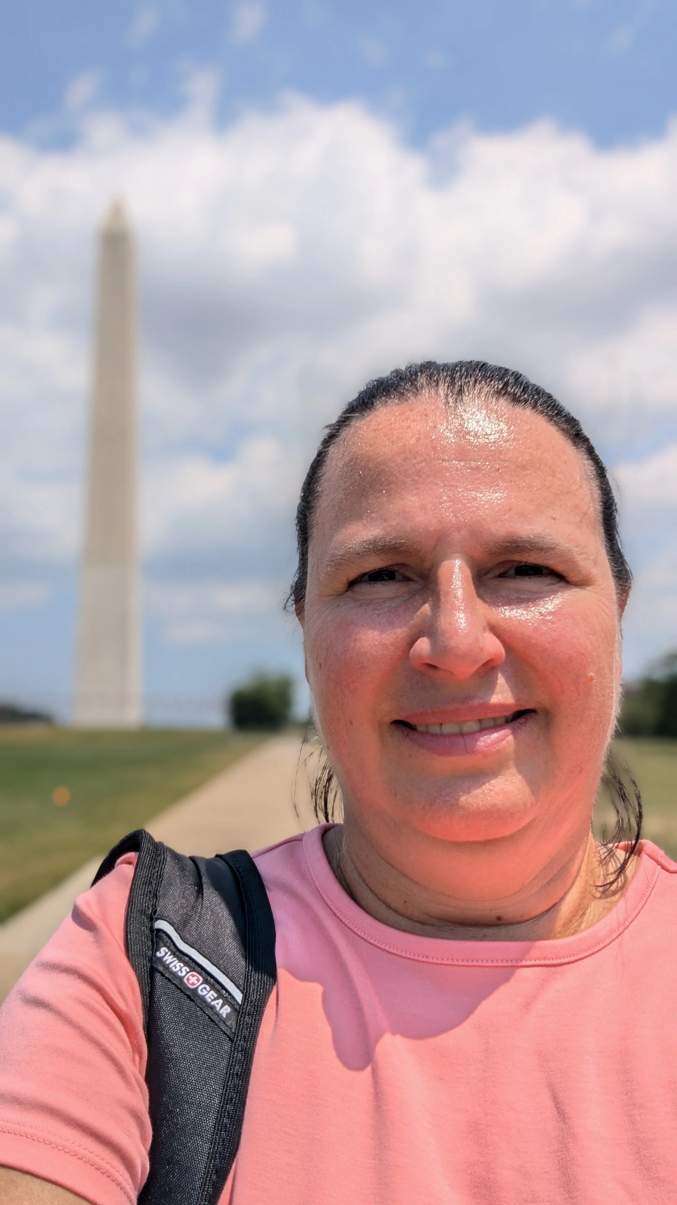 A woman with wet hair, smiling, wearing a pink shirt and backpack, taking a selfie with the Washington Monument in the background on a sunny day with scattered clouds.