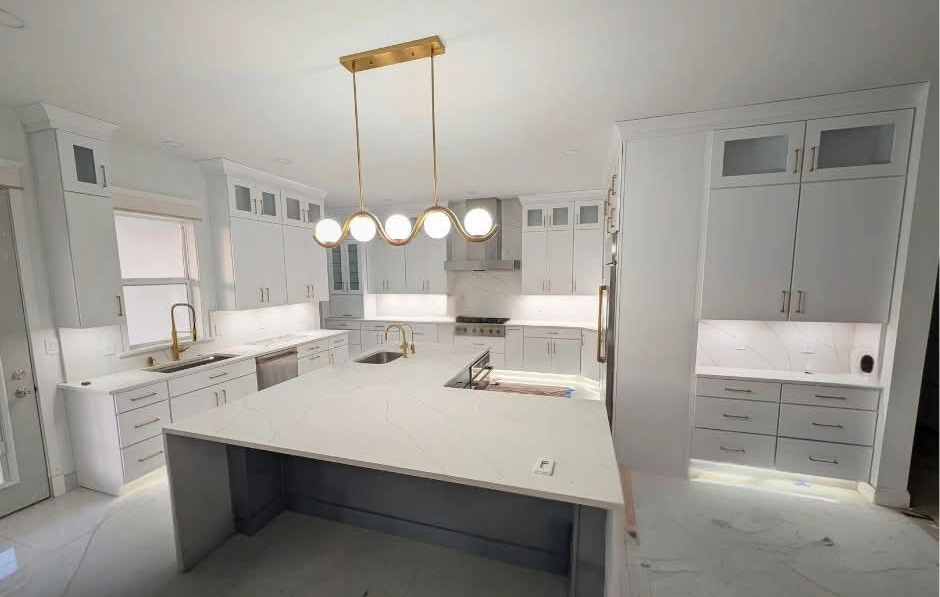 Modern white kitchen with gold fixtures, island countertop, and built-in cabinetry, appearance of under-cabinet lighting, and a ceiling chandelier.
