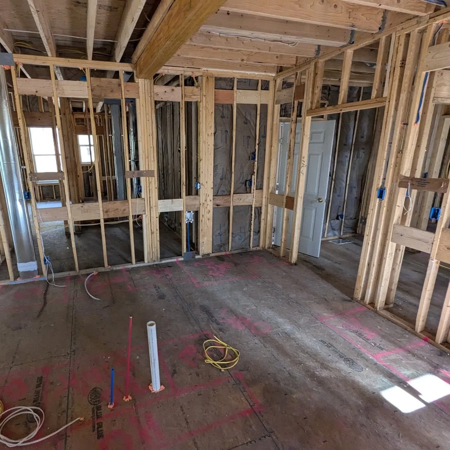 Interior view of a building under construction, showing exposed wooden framing, electrical wiring, pipes, and unfinished floors.