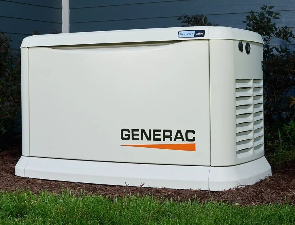 White Generac generator outside on grass and soil, with bushes and blue house siding in the background.