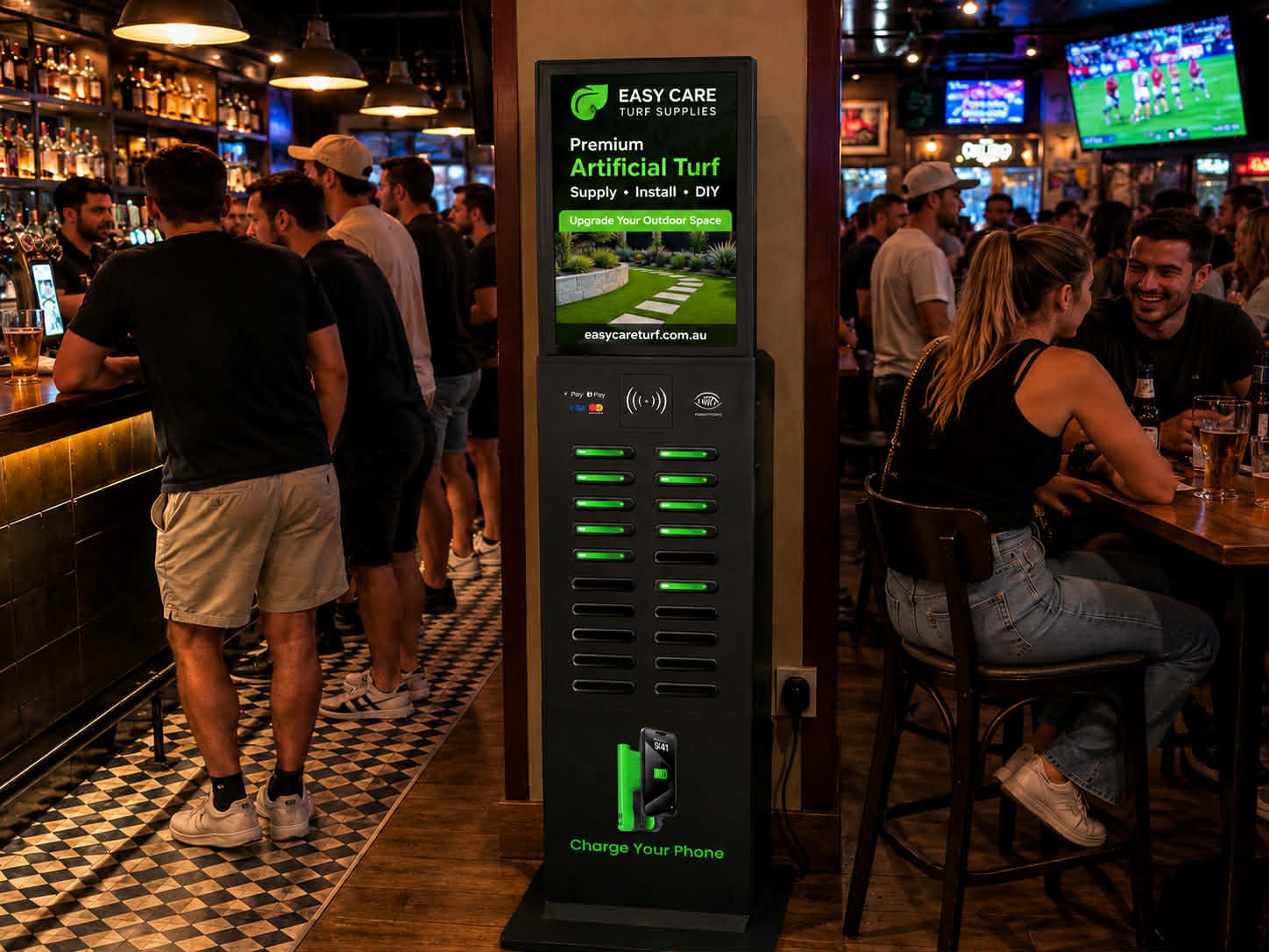 A crowded bar with people socializing and drinking, with a digital sign promoting artificial turf on the counter.