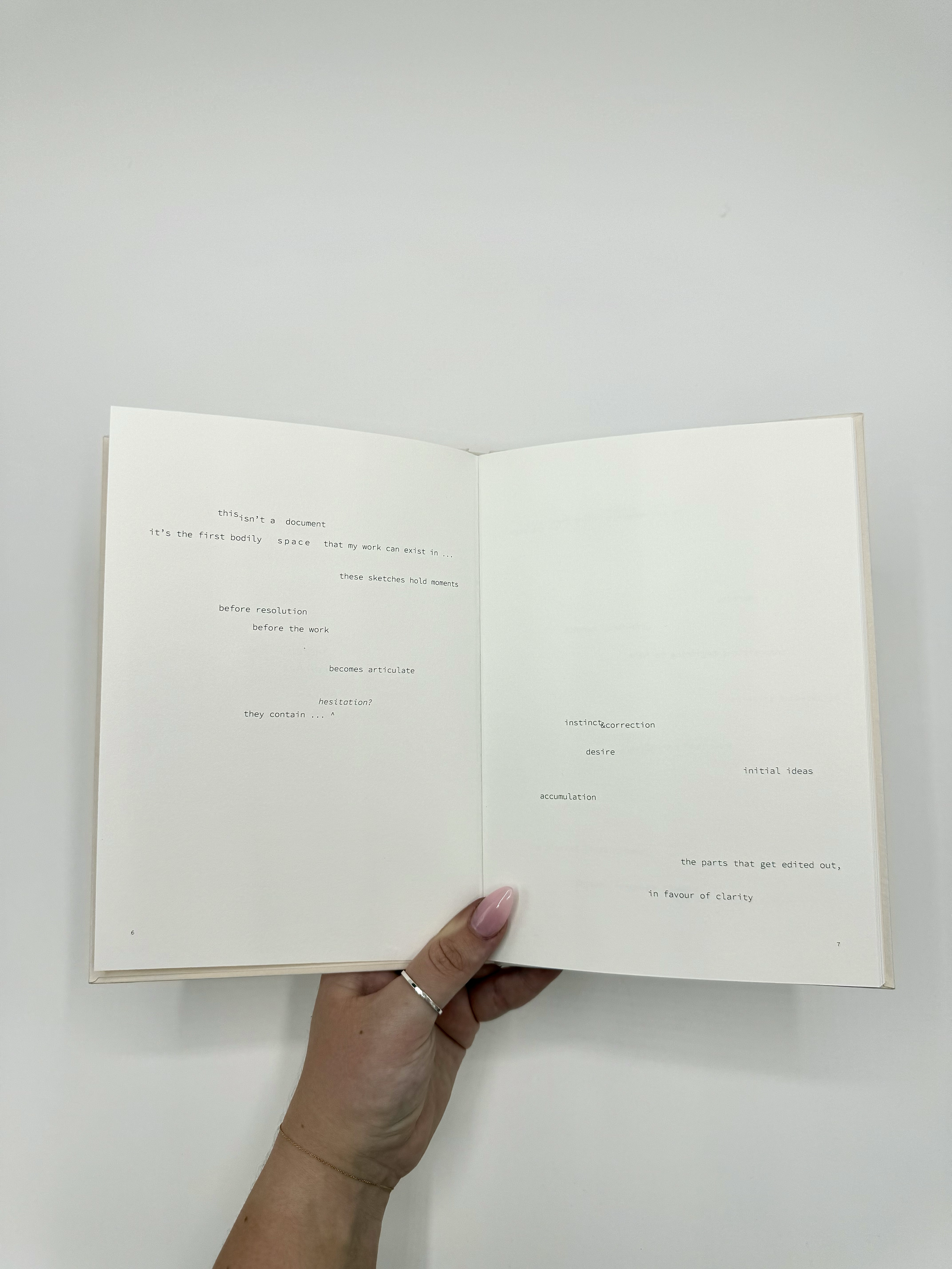 Open book being held by hand with a white background. The pages contain text in a minimalistic, typewriter style font with scattered words and phrases, mostly in lowercase.