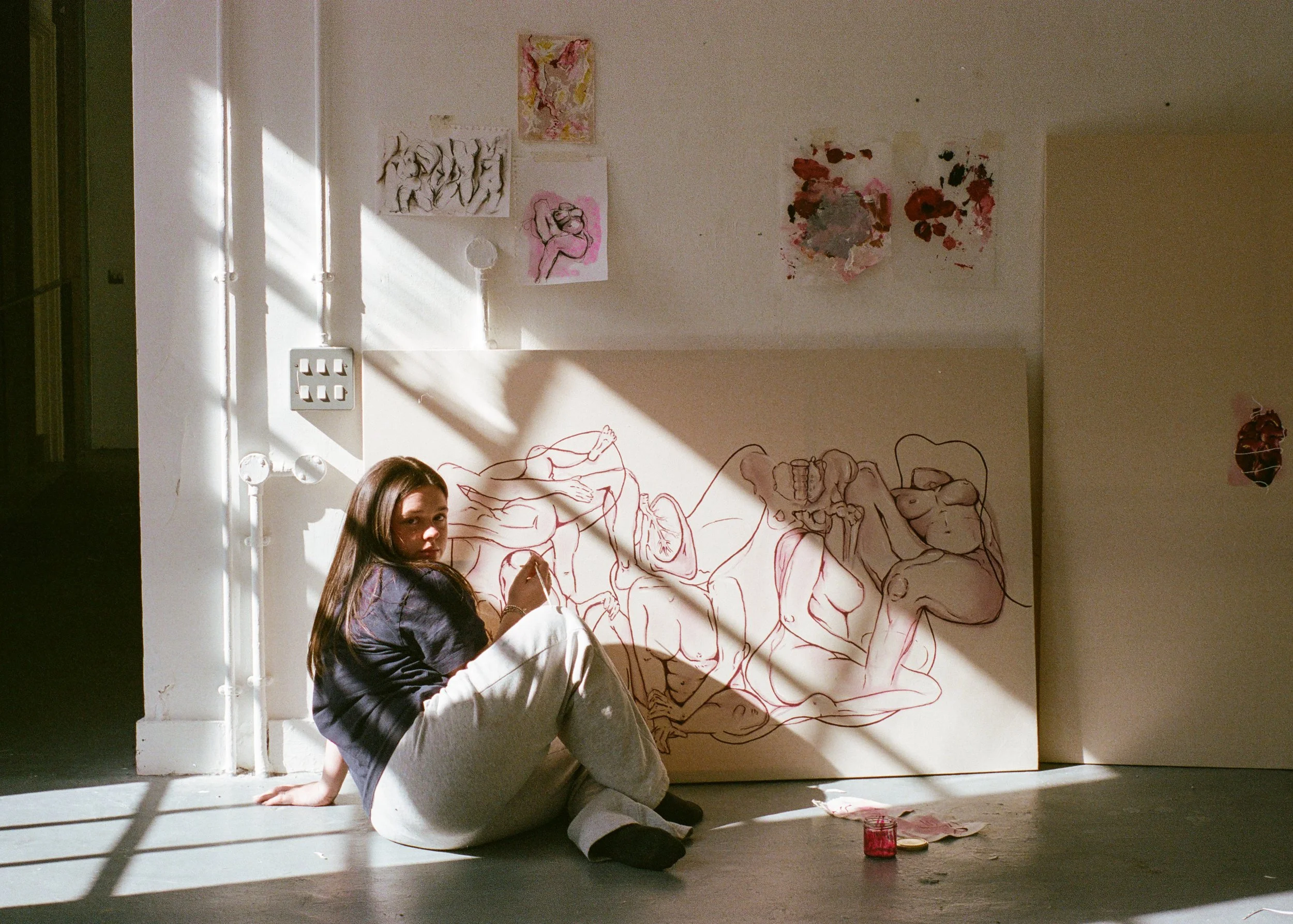 A young woman with long brown hair sitting on the floor in front of a wall adorned with various artworks. She is holding a paintbrush, and there are paint supplies on the floor nearby. The art on the wall includes sketches and paintings of human figures, some in color and some in outline.
