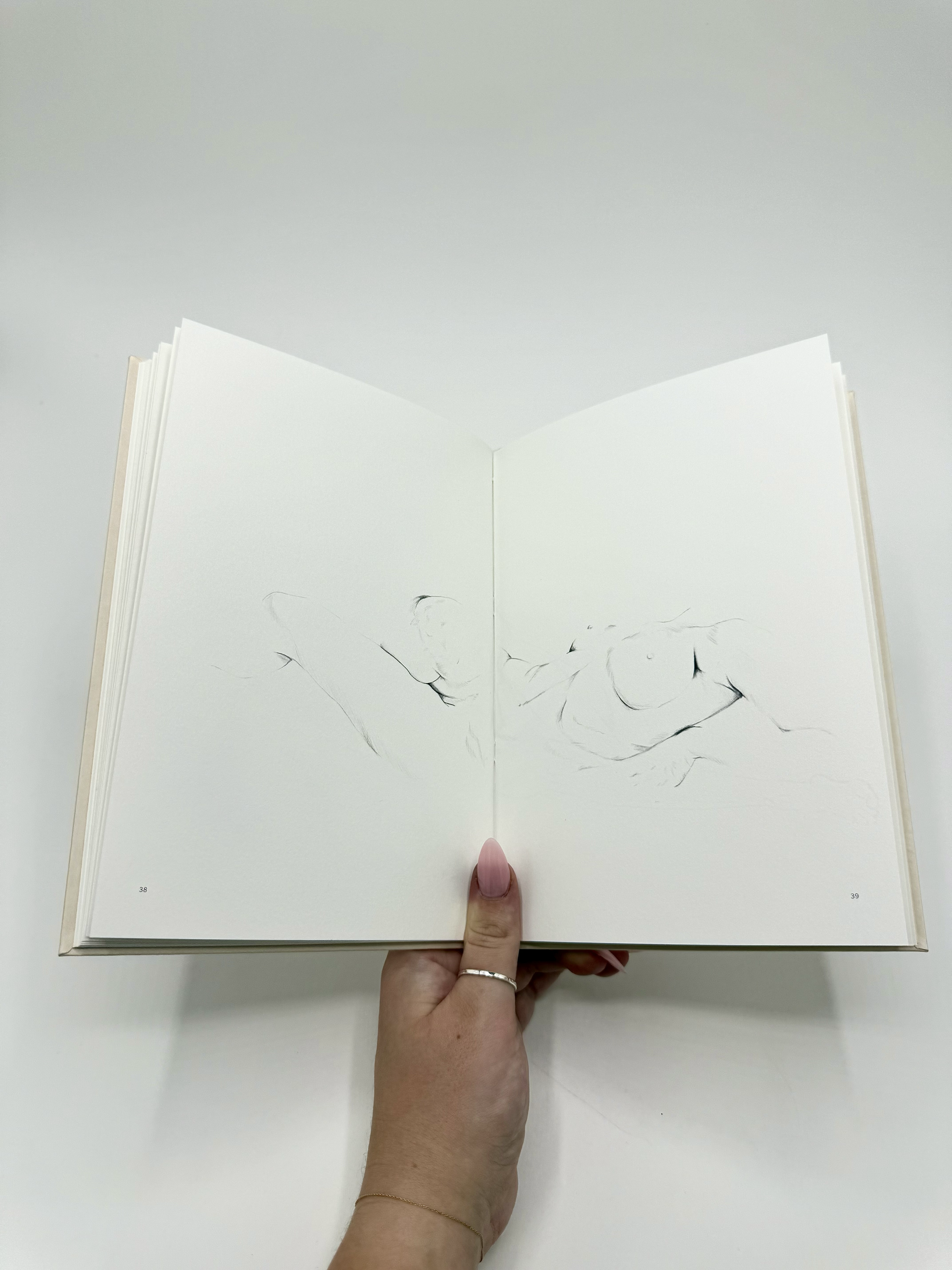 Open book with minimalist black line sketches of birds on pages 38 and 39, held by a person with long manicured nails, a ring, and a delicate bracelet, against a white background.