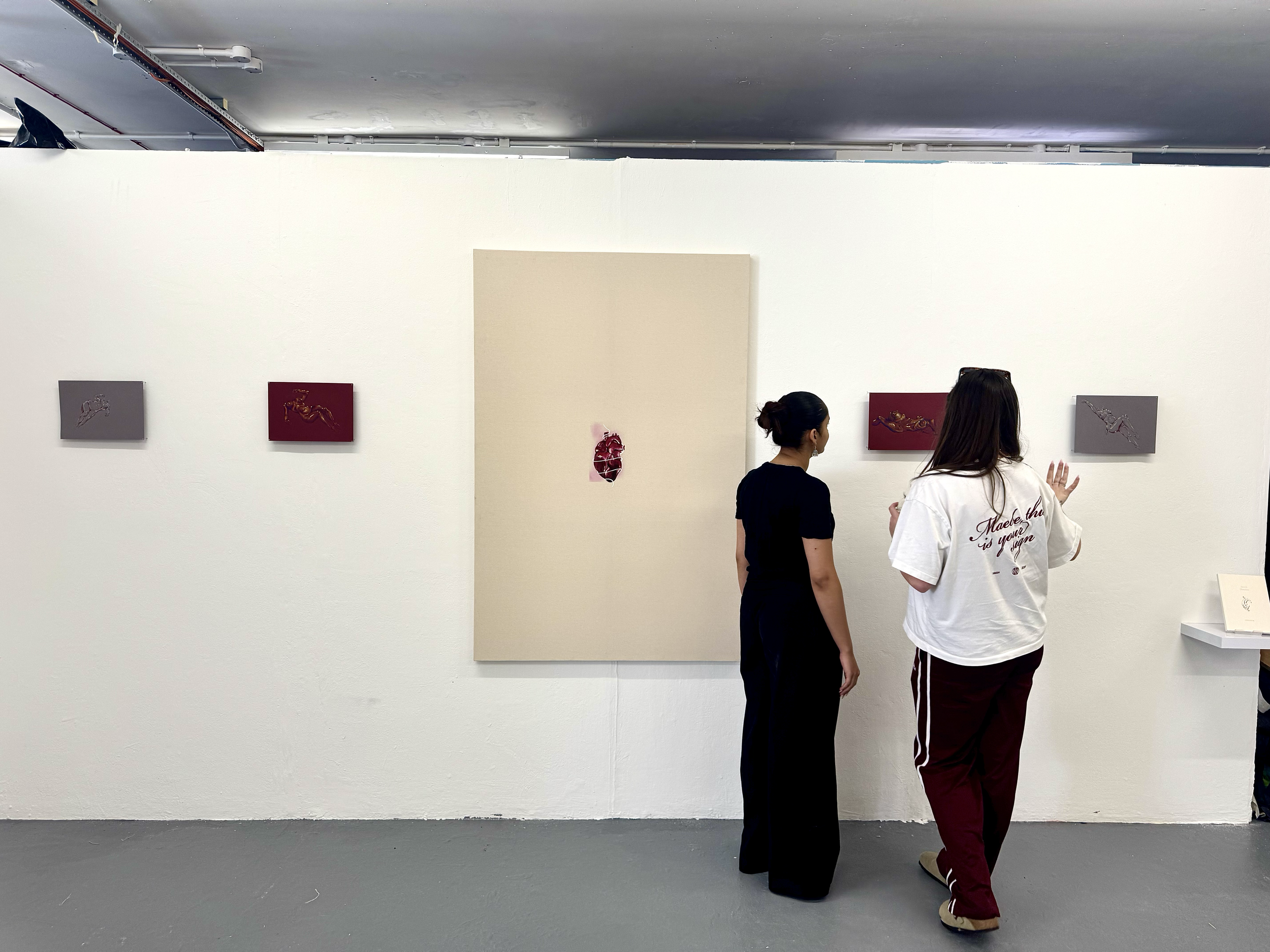 Two women standing in an art gallery, looking at paintings on the wall. The paintings are small, mostly dark-colored with some gold accents, and are mounted on gray and burgundy backgrounds.