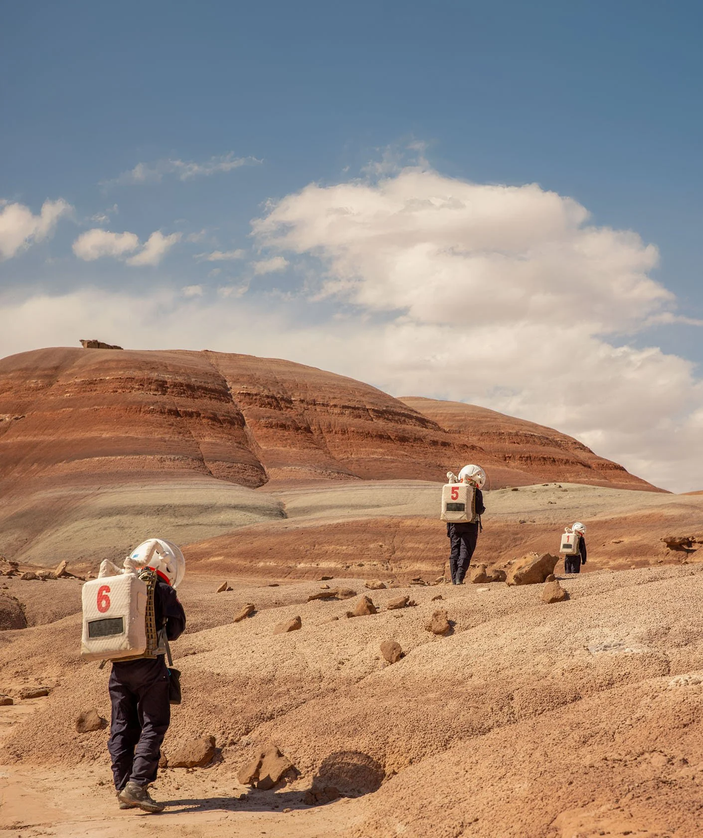 Four astronauts walking in a barren, rocky desert landscape with layered red and beige hills under a partly cloudy sky. Each astronaut has a numbered backpack, with visible numbers 6, 5, and 1.