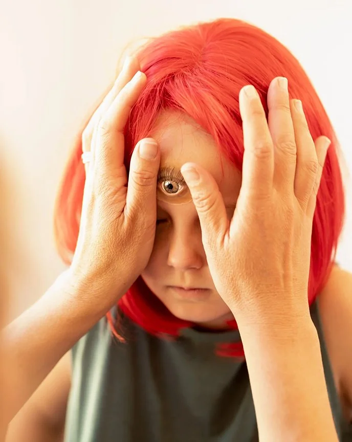A person with red hair looking through a small circular mirror held by their hands, focusing on their eye.