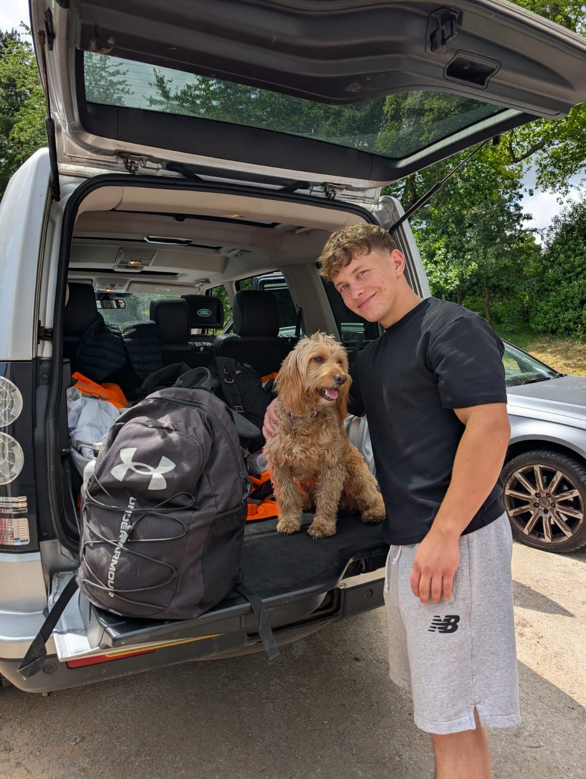 A young man standing next to a smiling golden doodle dog sitting in the open trunk of an SUV, which is filled with backpacks and outdoor gear, on a sunny day with trees in the background.