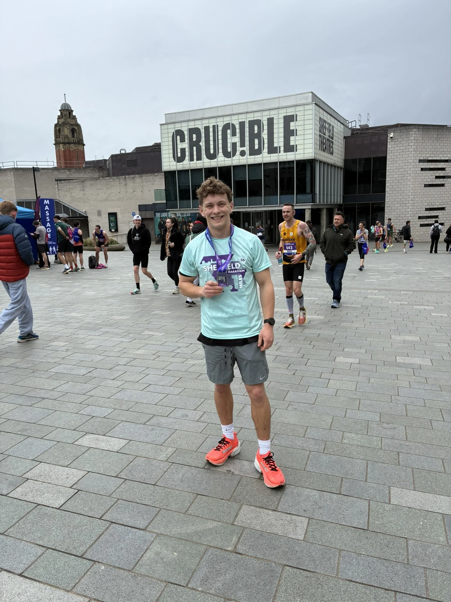 A young man smiling while holding a medal, standing in front of Sheffield Theatres, after completing a marathon.