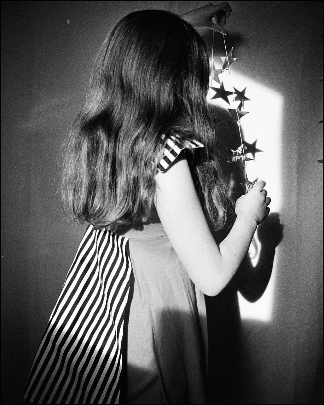 A girl with wavy hair is holding a string of star-shaped decorations near a light source, casting shadows on the wall.