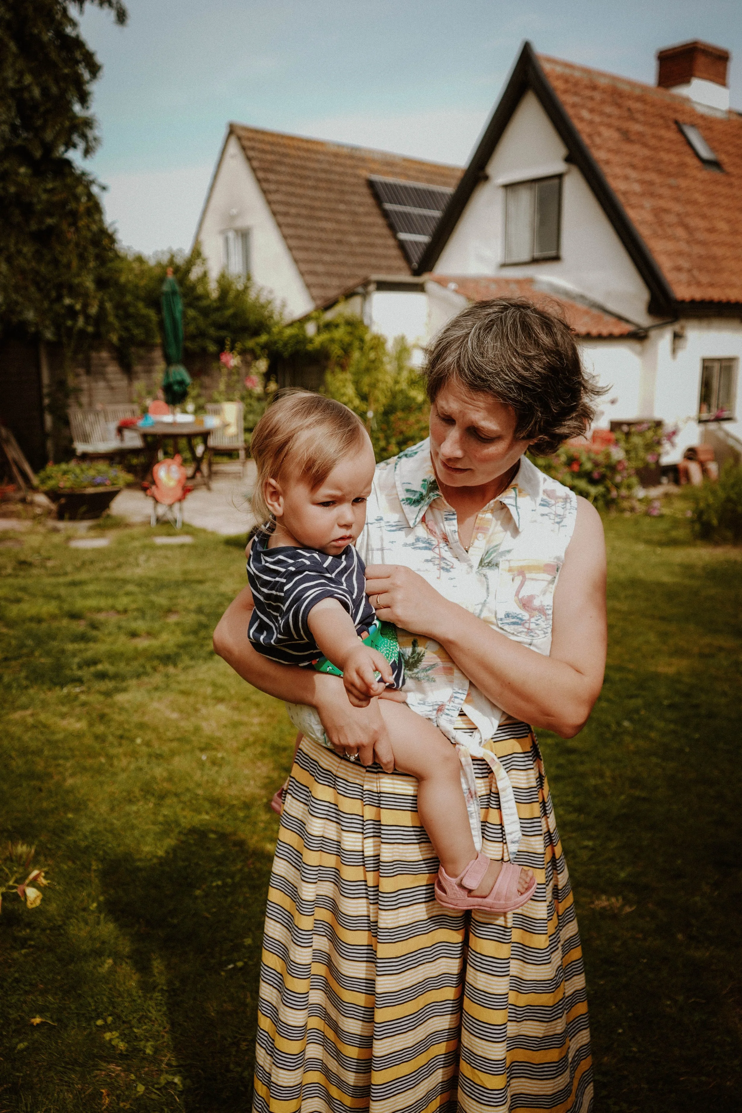 A woman holding a young girl in a backyard garden with a house in the background. The woman is wearing a striped skirt and sleeveless shirt, while the girl has on a striped navy and white shirt and pink shoes. The sky is clear, and the yard has patio