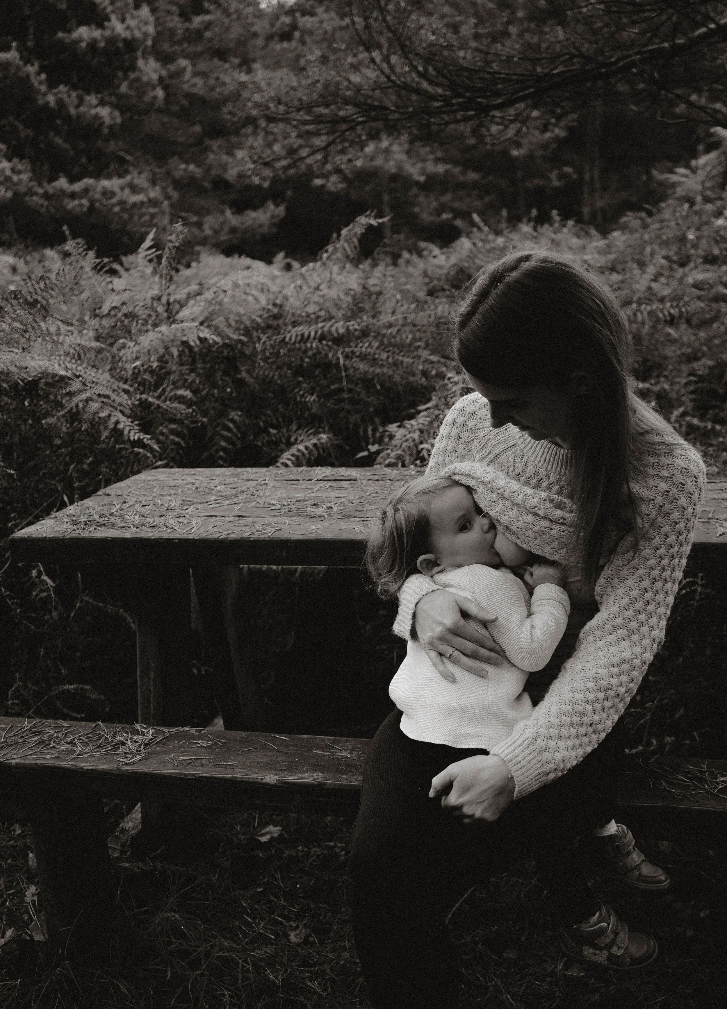 An adult woman with long hair and a child with curly hair sitting on a bench outdoors amidst ferns and trees, the woman appears to be kissing and hugging the child, and both are dressed in cozy sweaters.