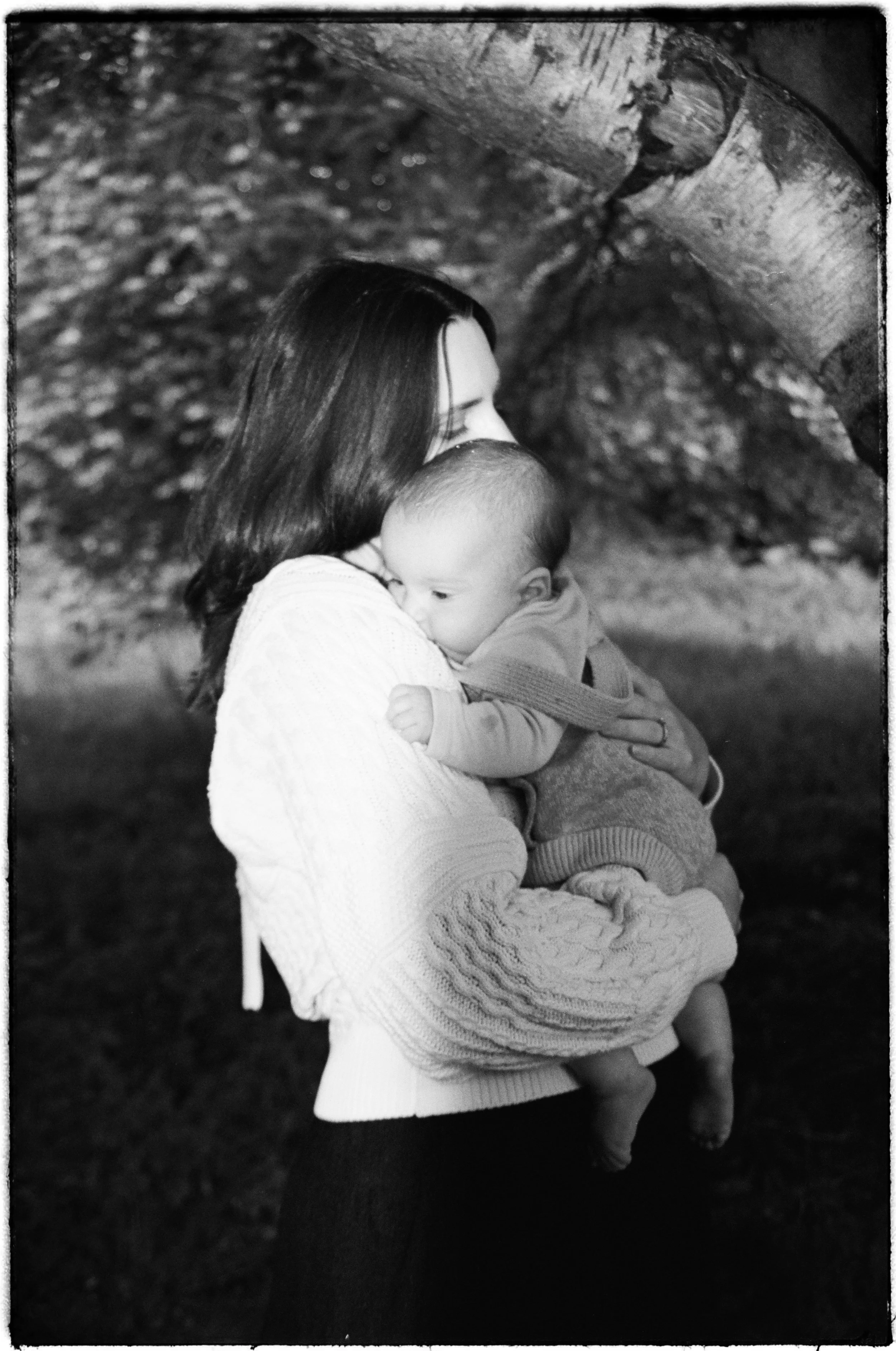 A woman with dark hair holding a baby close, both facing each other in an outdoor setting. The image is in black and white.