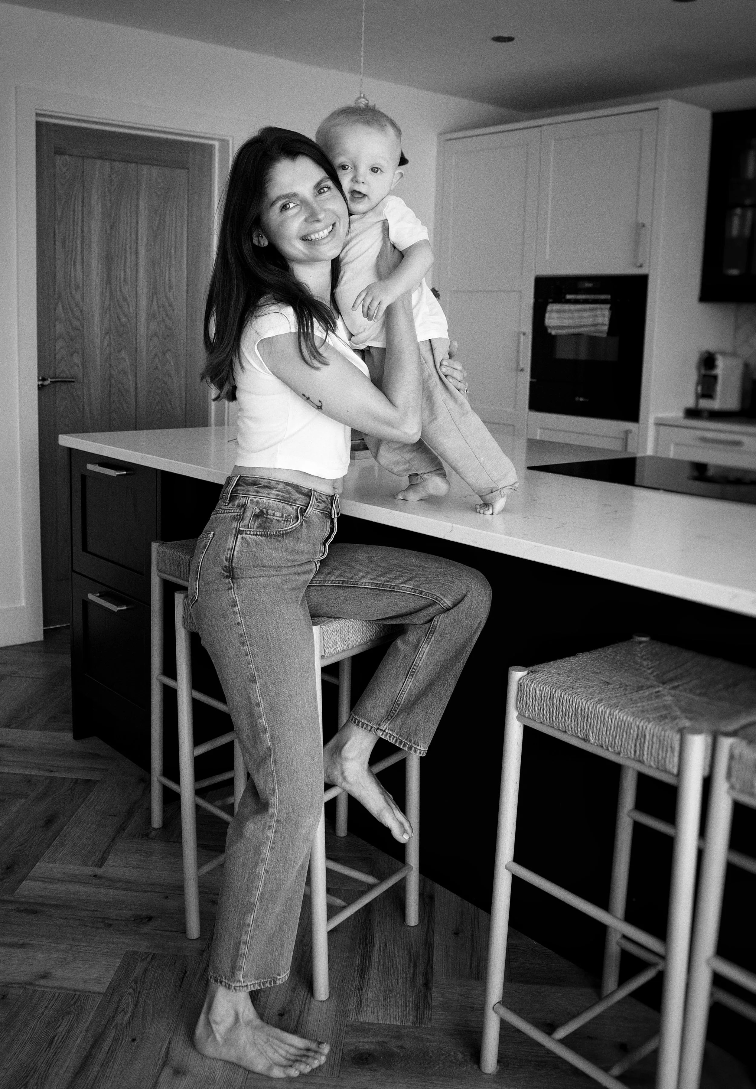 A woman with dark hair smiling and holding a toddler in a kitchen. The woman is sitting barefoot on a barstool, with her right foot resting on the ground. The toddler is standing on a kitchen counter, leaning on the woman, with a happy expression and