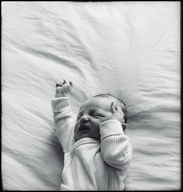 Black and white photo of a sleeping baby lying on a soft surface with one arm raised and a hand near the head.