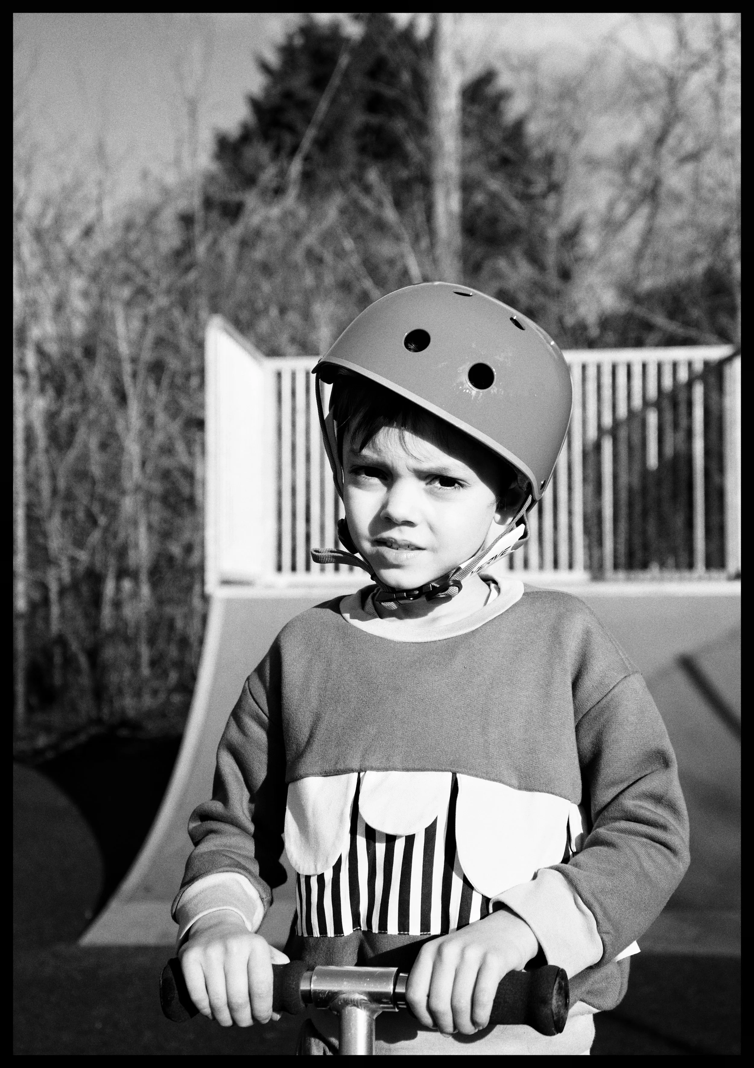 A young boy wearing a helmet rides a scooter outdoors, with trees and a wooden fence in the background.