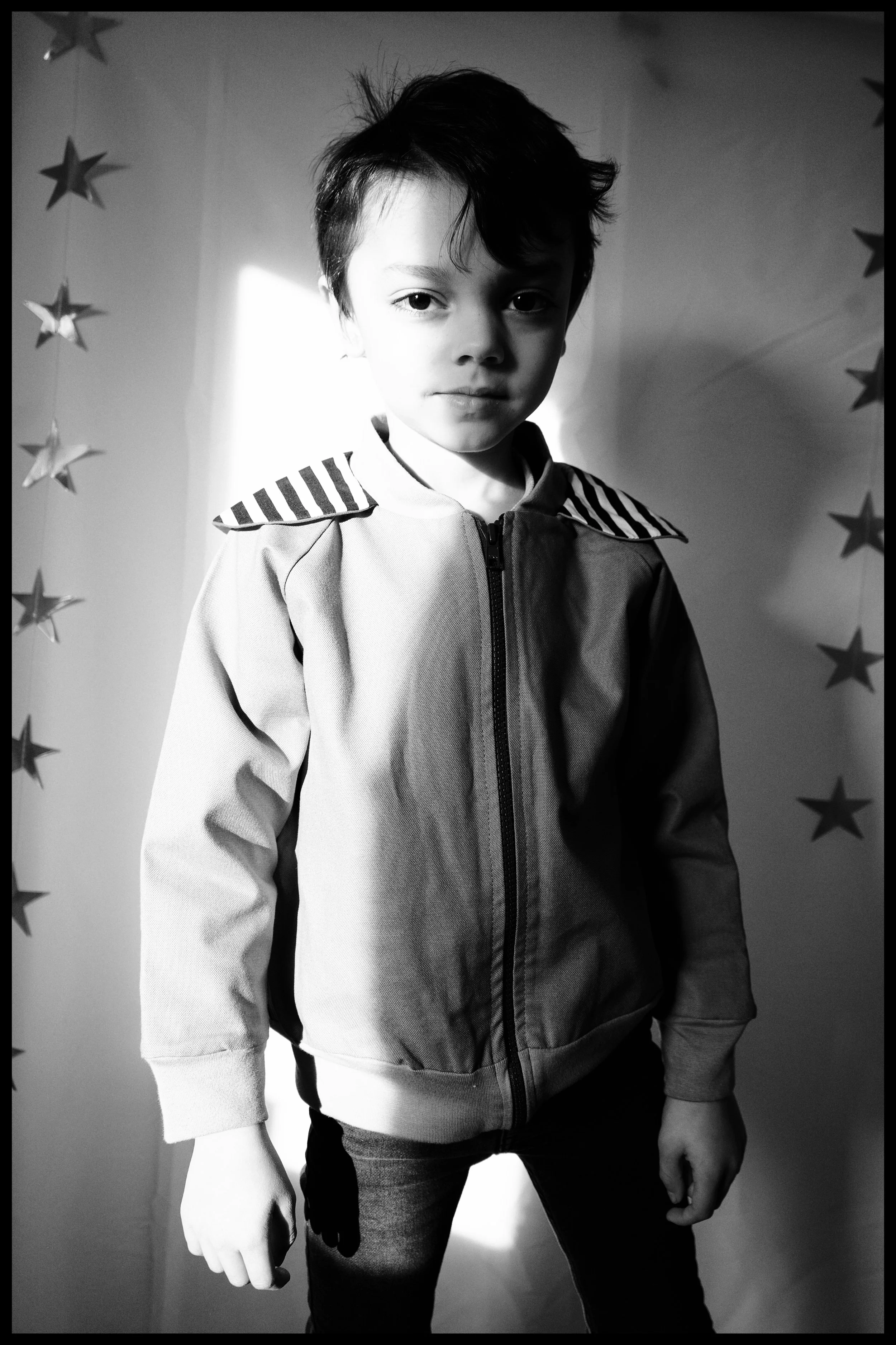 A young boy with dark hair, wearing a zip-up jacket, standing in front of a backdrop decorated with hanging star shapes. The photo is in black and white, and the boy has a serious expression.