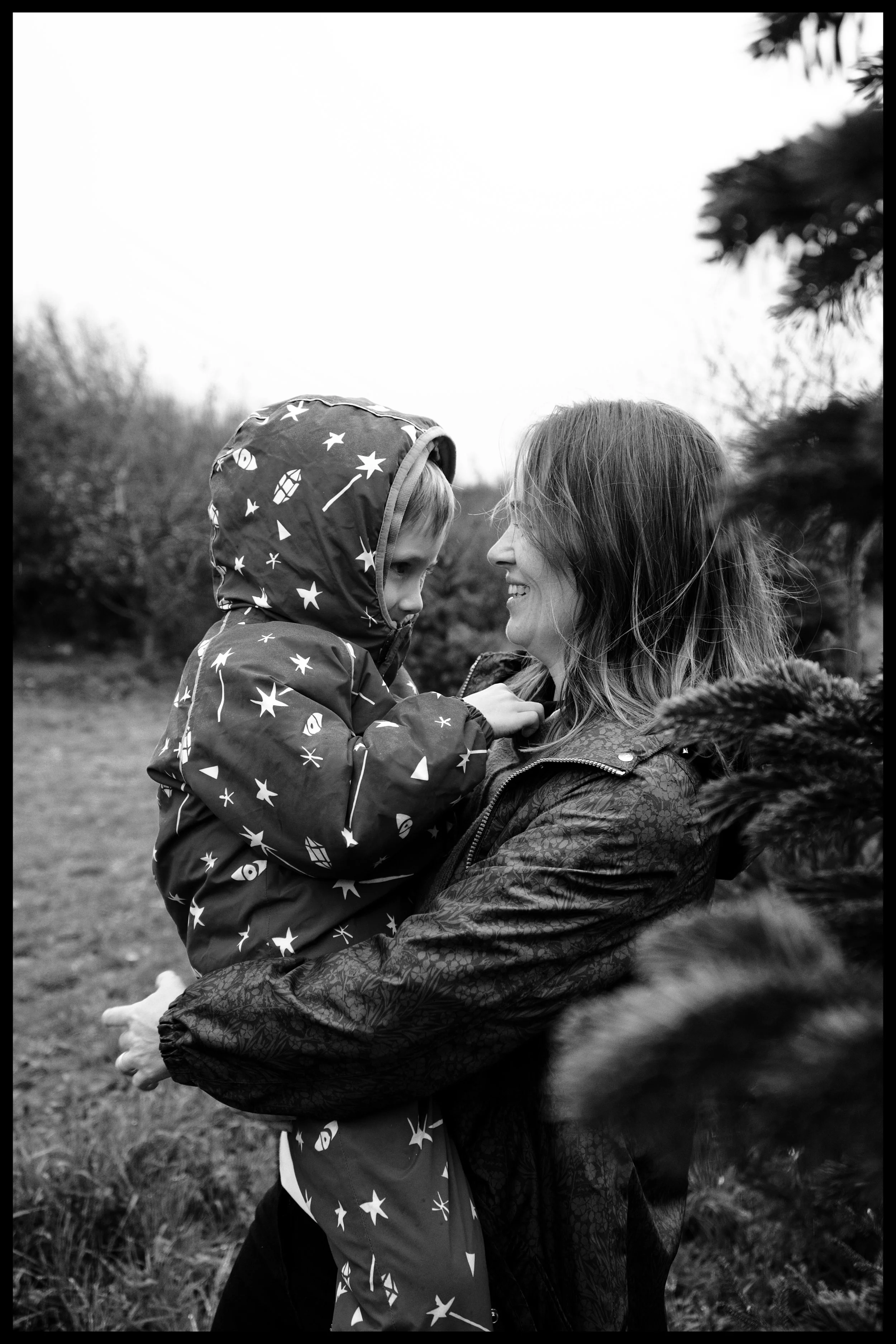 A woman and a young girl are outside, smiling and looking at each other. The woman is wearing a patterned jacket, and the girl is wearing a jacket with star and rocket patterns. They are holding each other's hands, with the woman lifting the girl sli