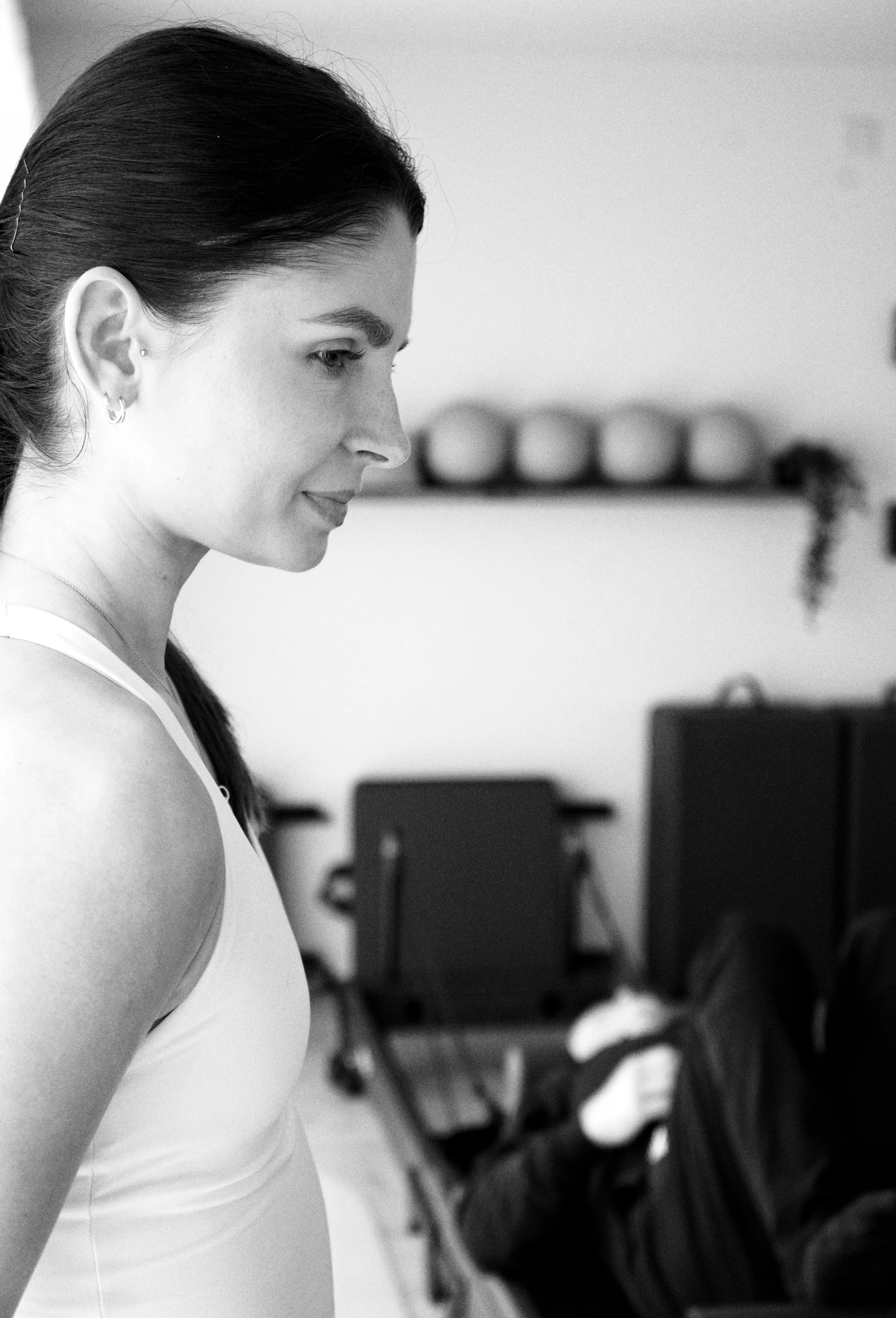 A woman with dark hair tied back, wearing a white sleeveless top and earrings, is smiling while looking down. In the blurred background, there are ballet shoes on a shelf and dance studio furniture.