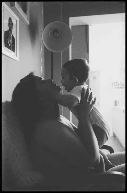 A woman holding a young child close to her face, both smiling, indoors with framed photos on the wall and a doorway in the background.