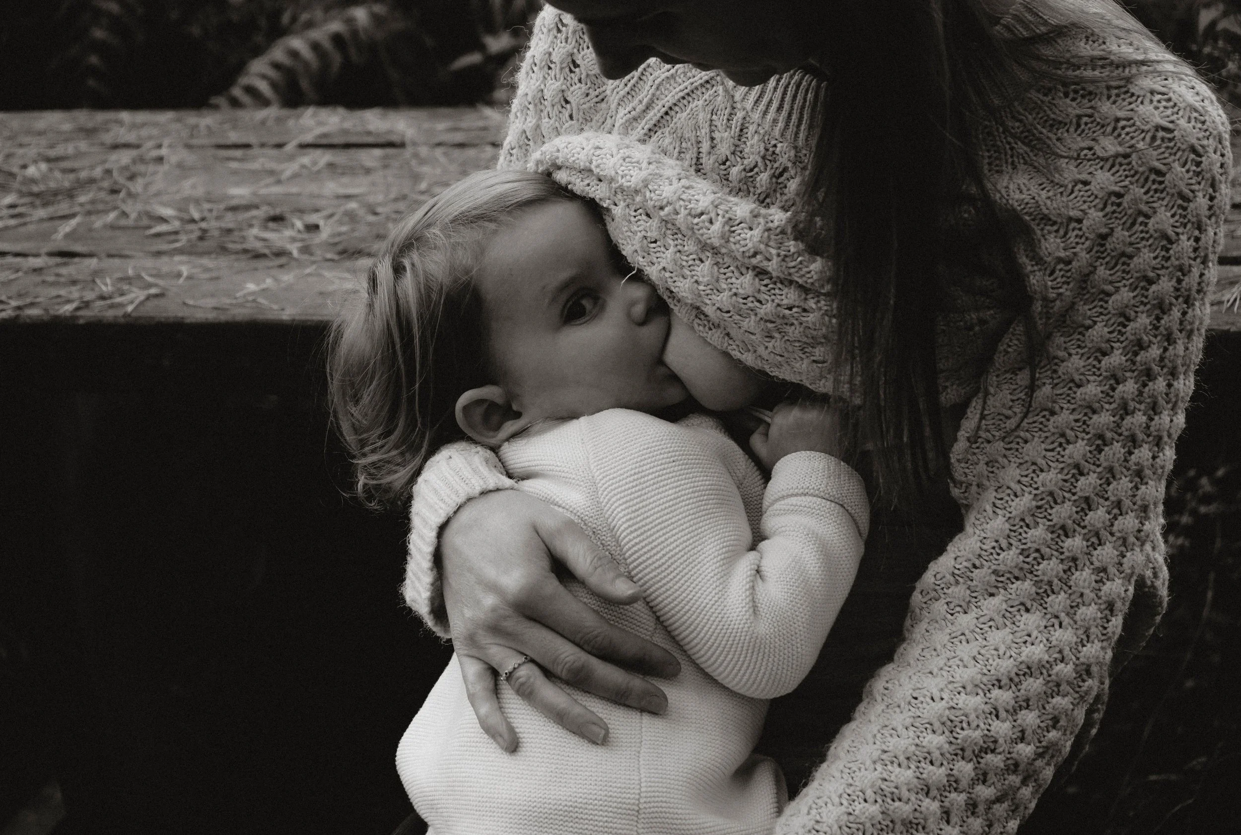A woman with long dark hair wearing a chunky knit sweater kisses a young girl on the cheek. The girl has blonde hair, is wearing a light-colored sweater, and is holding her thumb near her mouth while looking away from the camera. The setting appears 