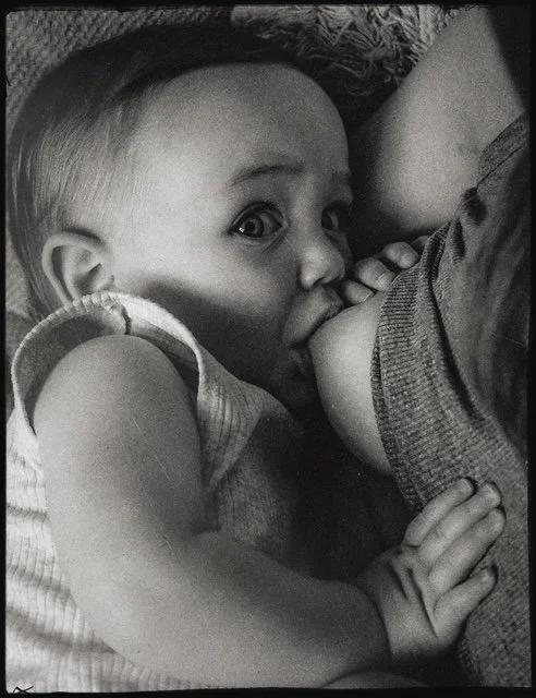 A young childlying on the floor, breastfeeding, and looking toward the camera.