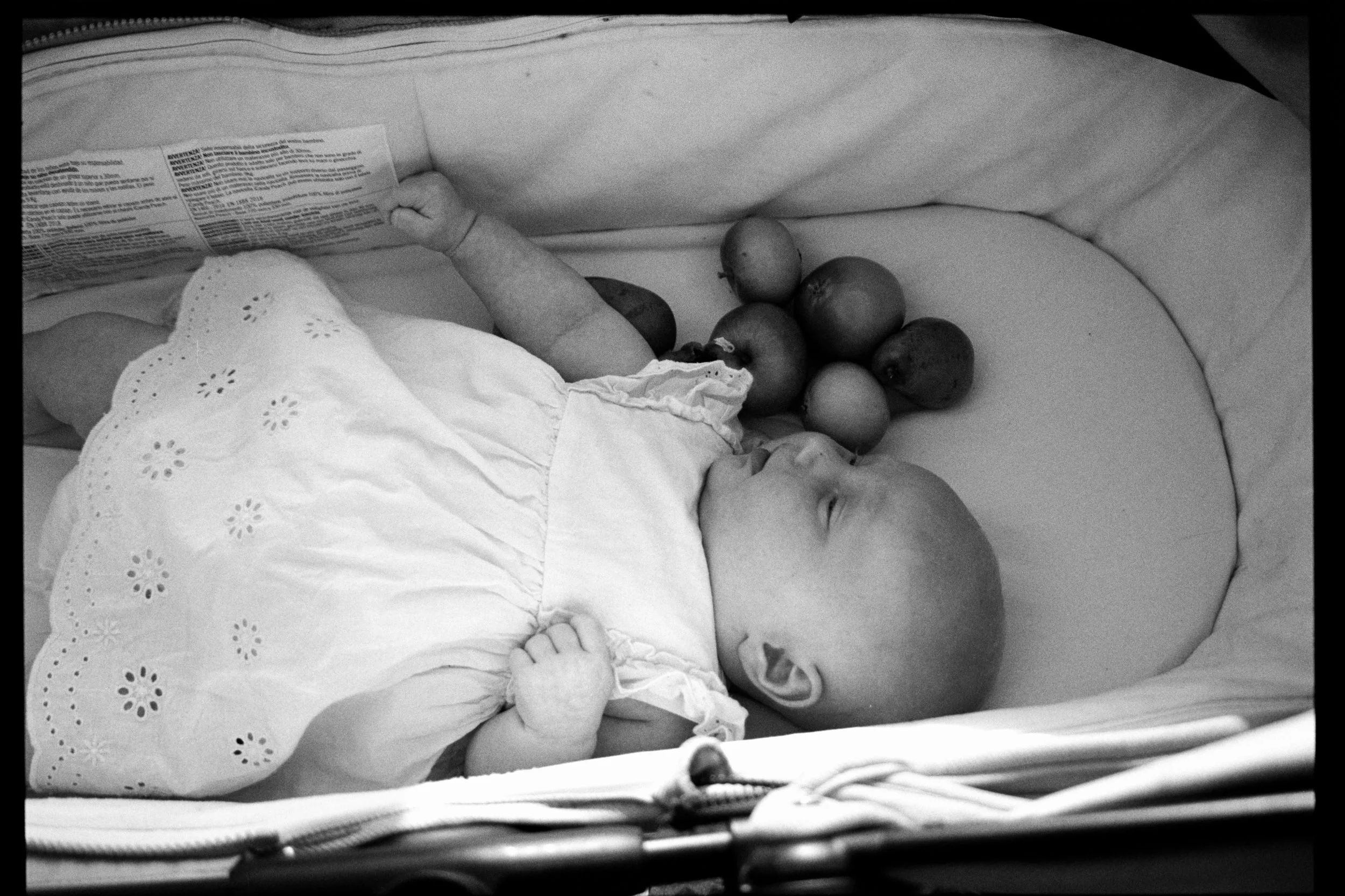A baby lying on its back in a crib, wearing a white dress with eyelet embroidery, holding a small object, with a bunch of apples placed on the mattress nearby, and a package with text partially visible.
