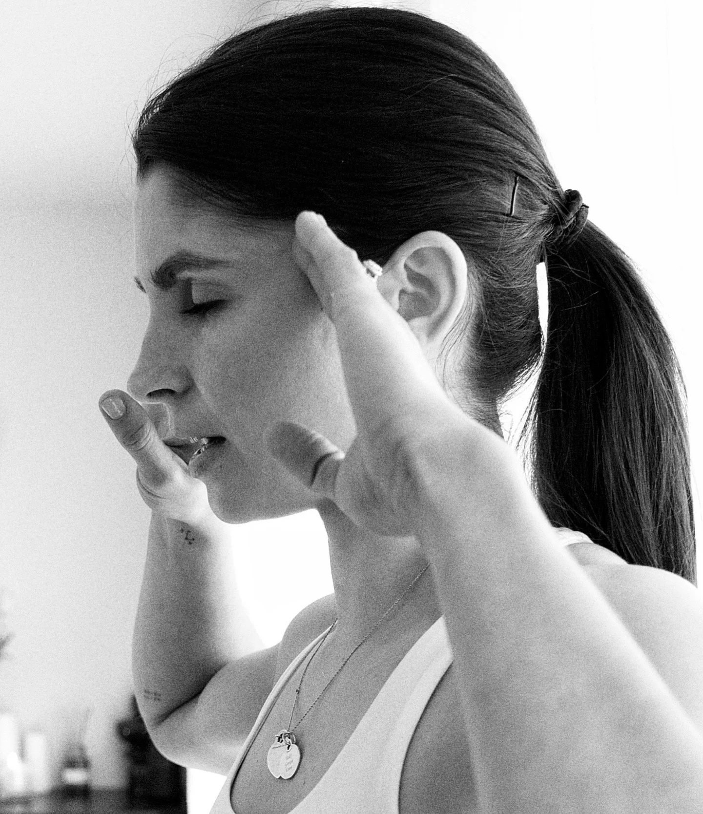 A woman with long dark hair pulled back in a ponytail, eyes closed, resting her head in her hand, wearing a sleeveless top, necklace with a round pendant, and a ring on her finger.
