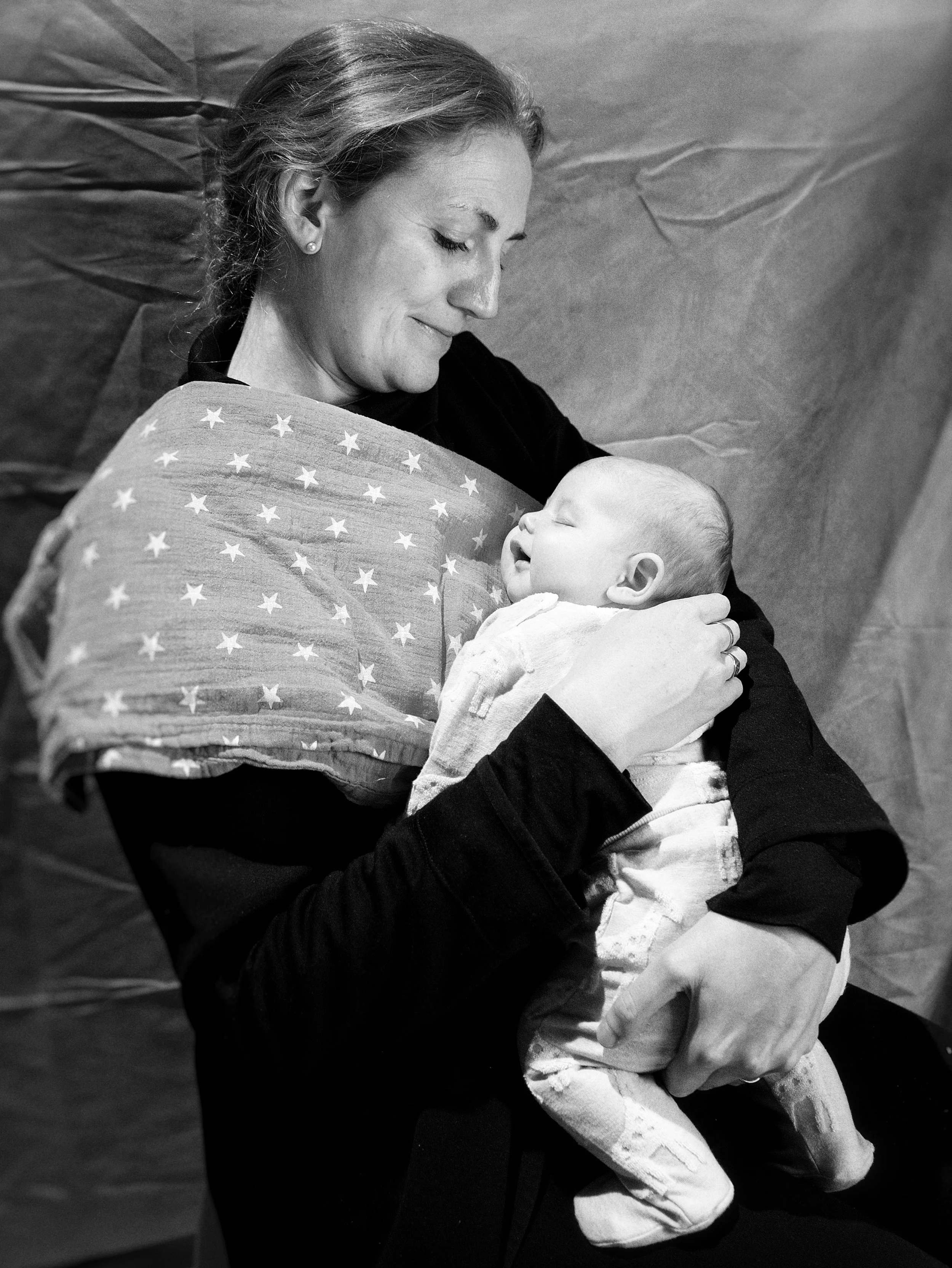 A woman with light-colored hair smiling and holding a baby dressed in light-colored clothing, with the woman wearing earrings and a sweater with a star pattern, in black and white.