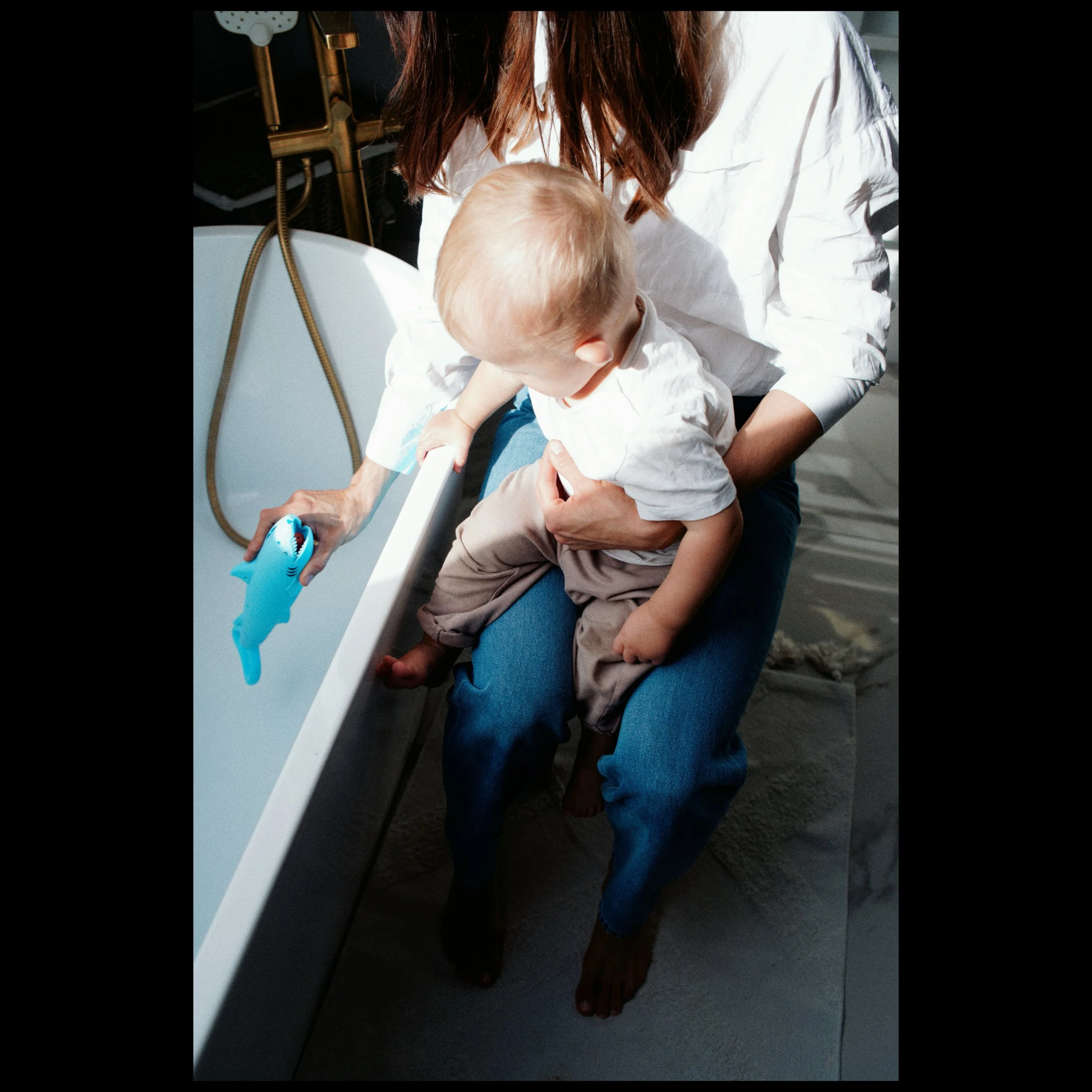 A person with red hair and light skin holding a young child, with the child's hand extended into a blue toy set, near a white infant bathtub on a dark floor.