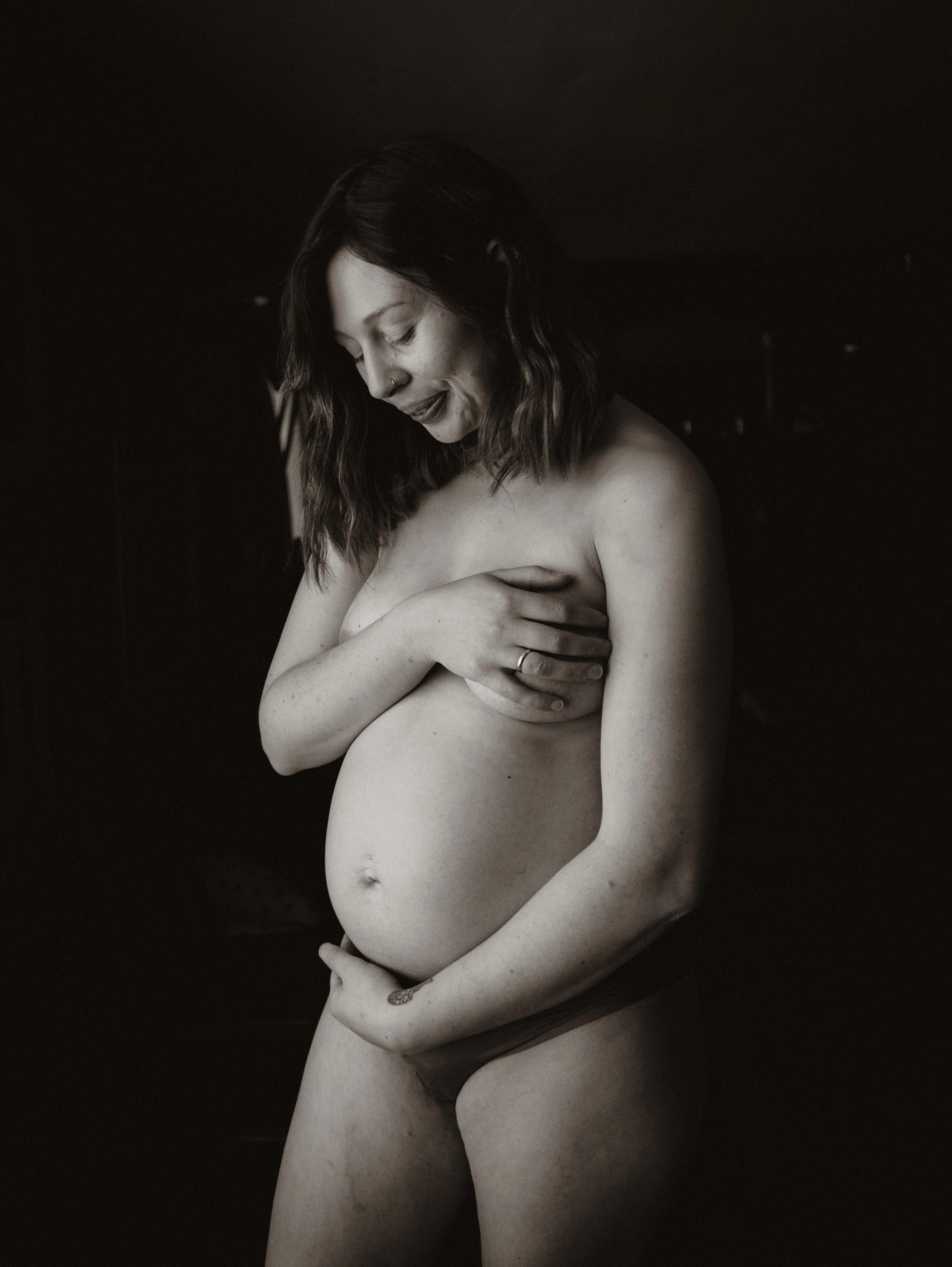 Black and white photograph of a pregnant woman with long wavy hair, holding her breasts and looking downward, with her belly prominently displayed.