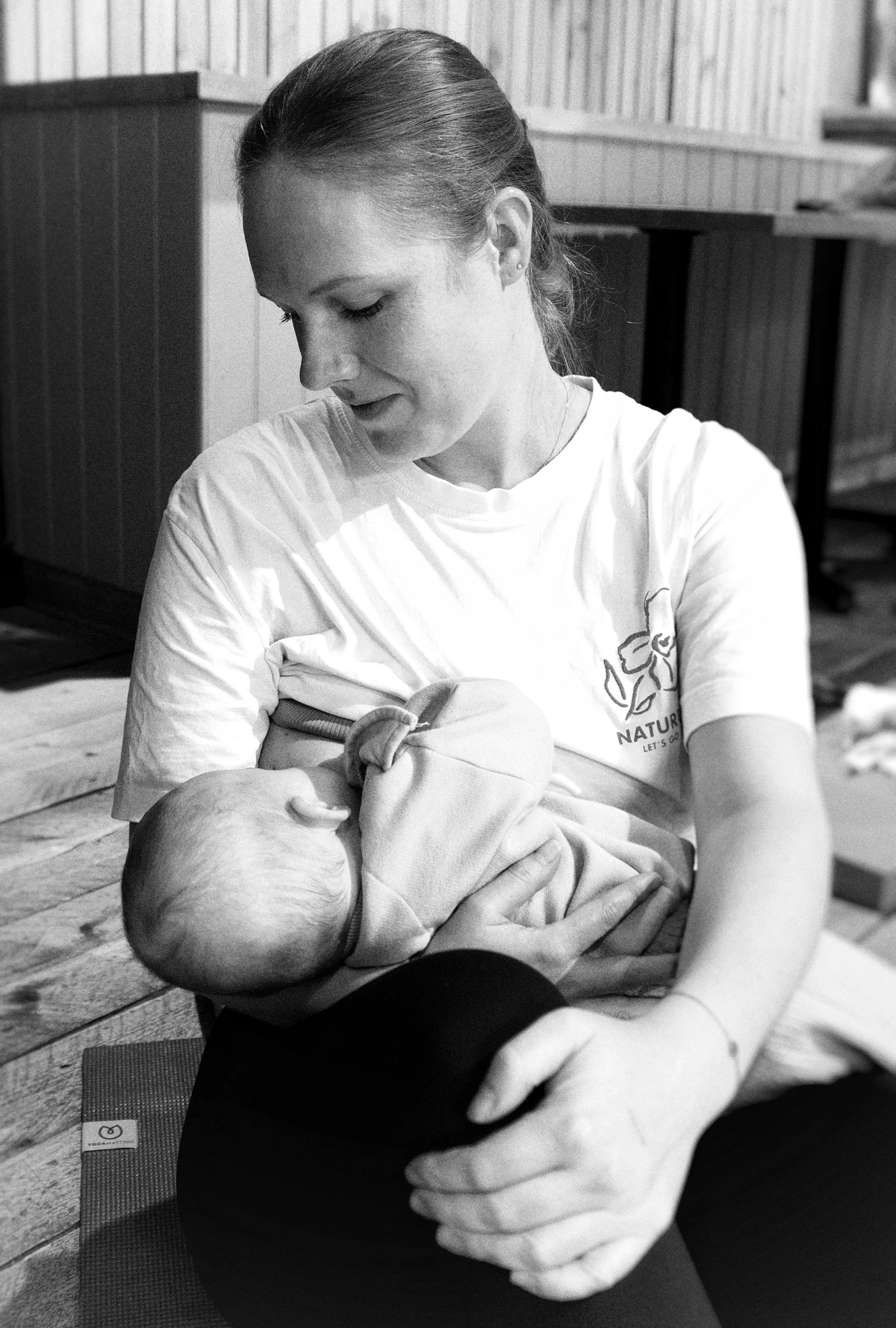 A woman in a white t-shirt with a logo is practicing yoga, holding a baby in her arms in a room with wooden floors and walls.