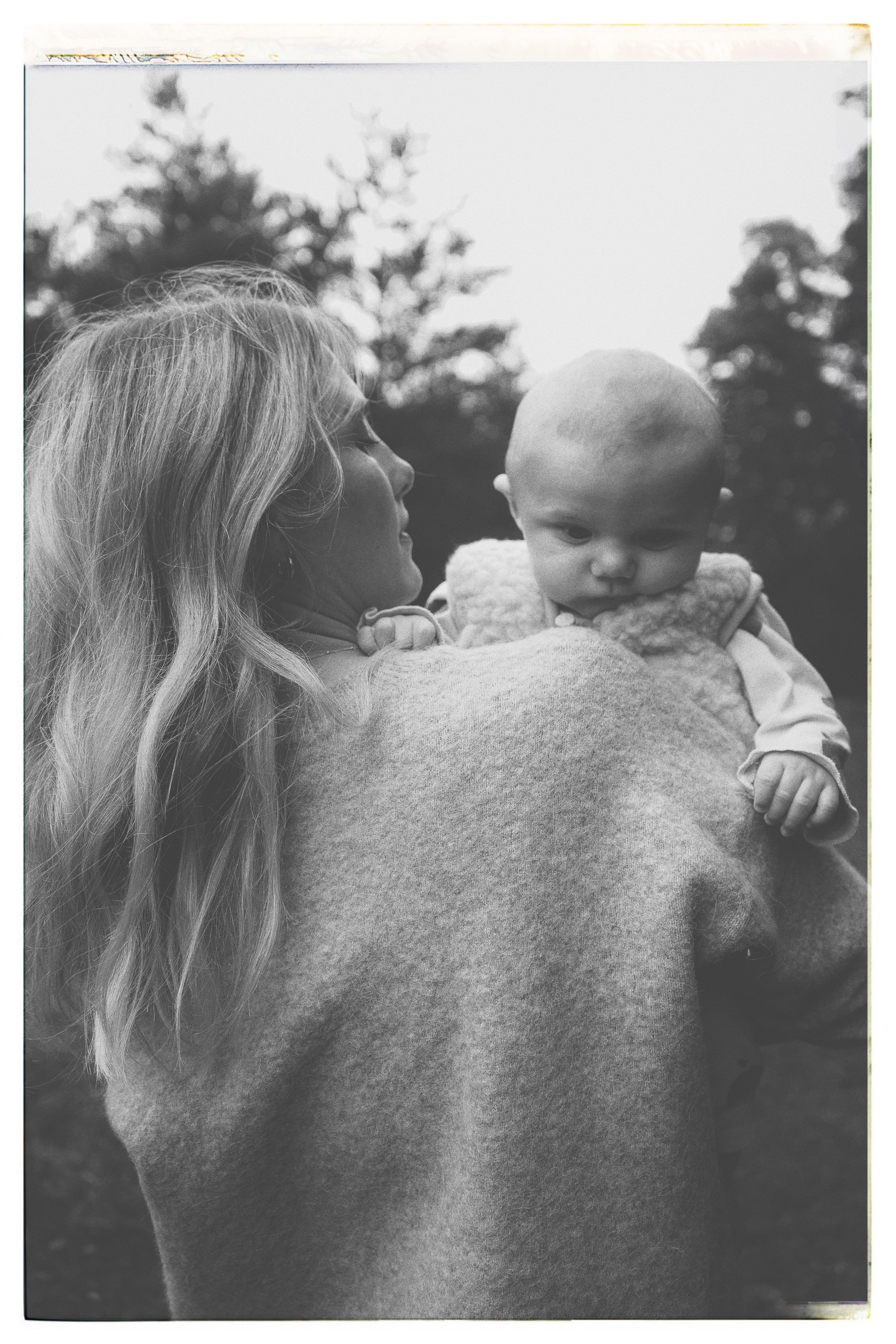 A woman with long wavy hair is facing right, holding a baby with a shaved head and chubby cheeks, gazing downward, outdoors with blurred trees in the background.