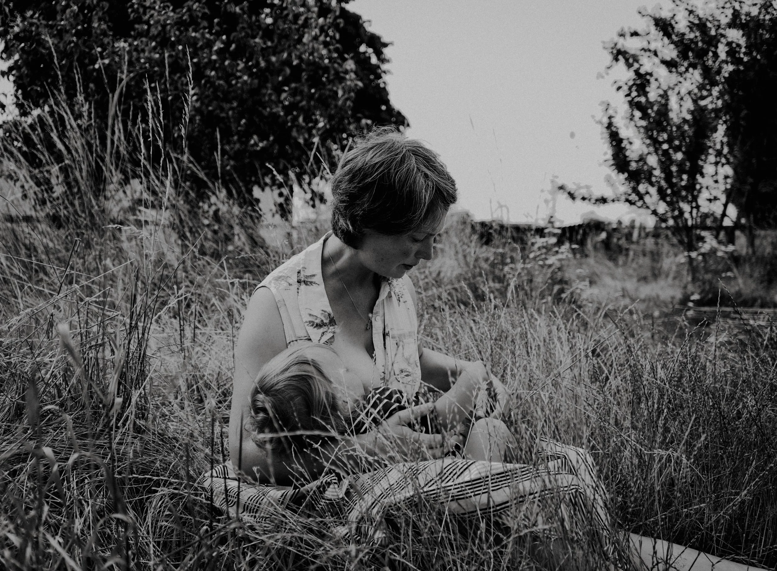 A woman with short hair sitting in a field of tall grass, looking down, with trees in the background, in black and white.