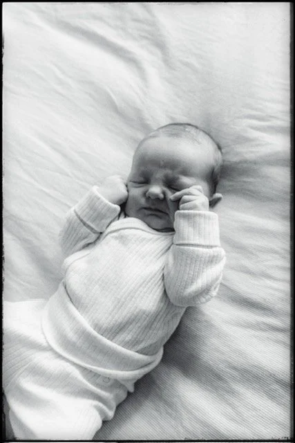 Black and white photo of a newborn baby sleeping on a soft surface, wearing a long-sleeve shirt and pants.