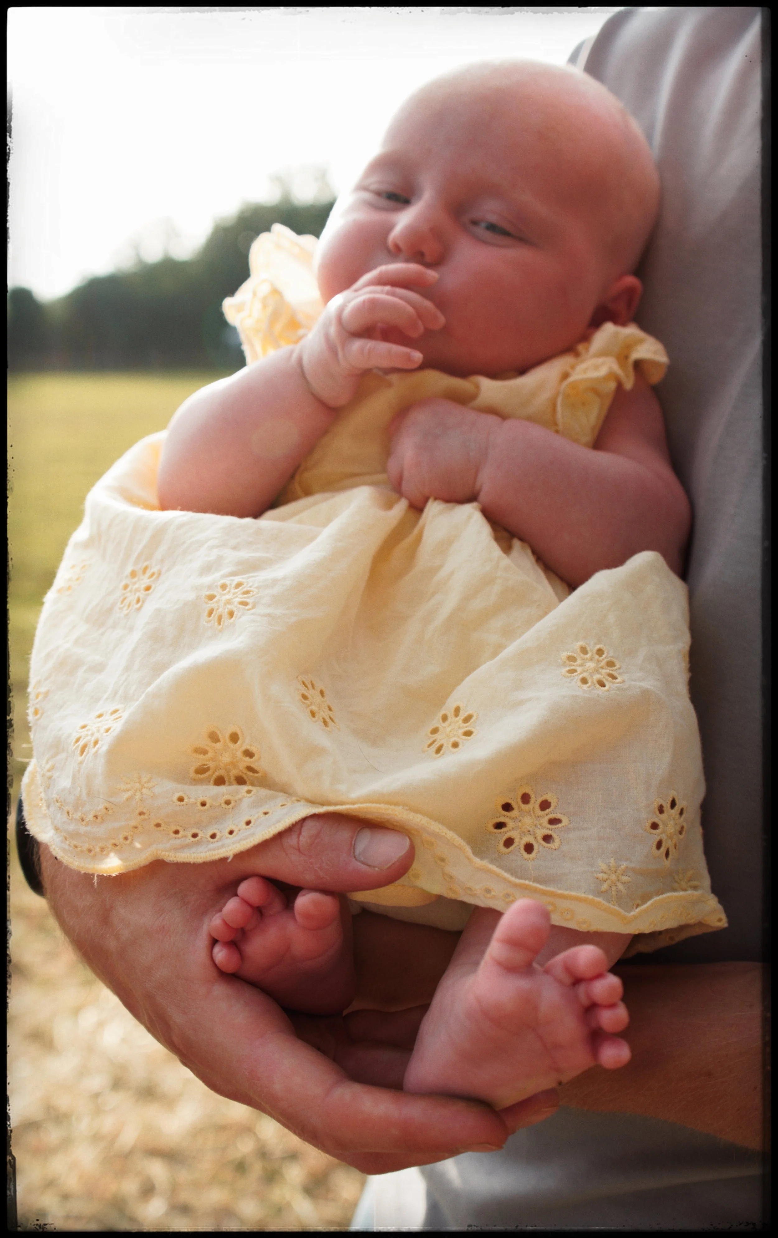 A baby dressed in a yellow embroidered dress being held by an adult outdoors, with a blurred natural background.