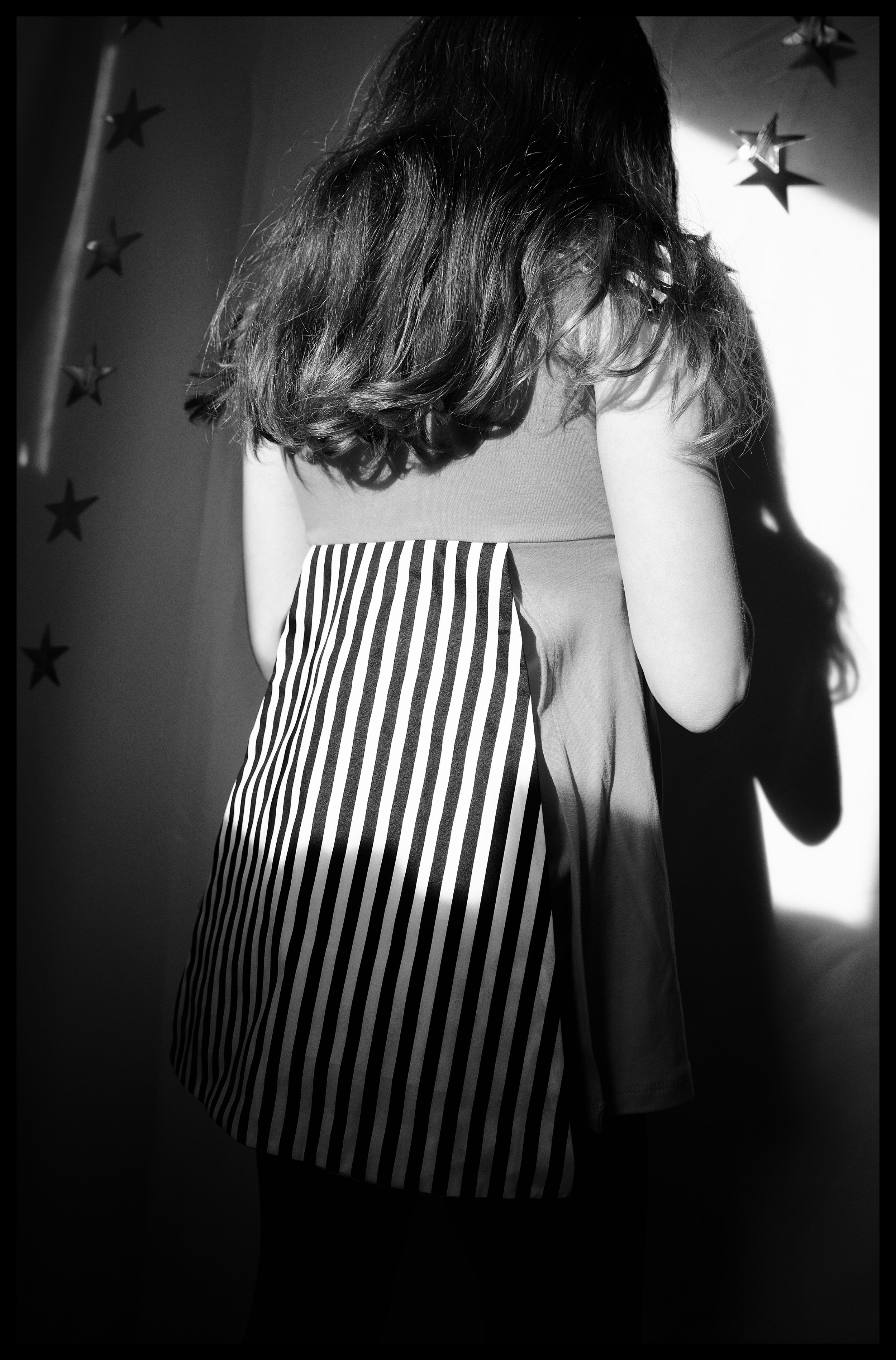 Black and white photo of a woman with long hair, wearing a top and a striped skirt, standing with her back to the camera in a dimly lit room decorated with star-shaped objects on the wall.