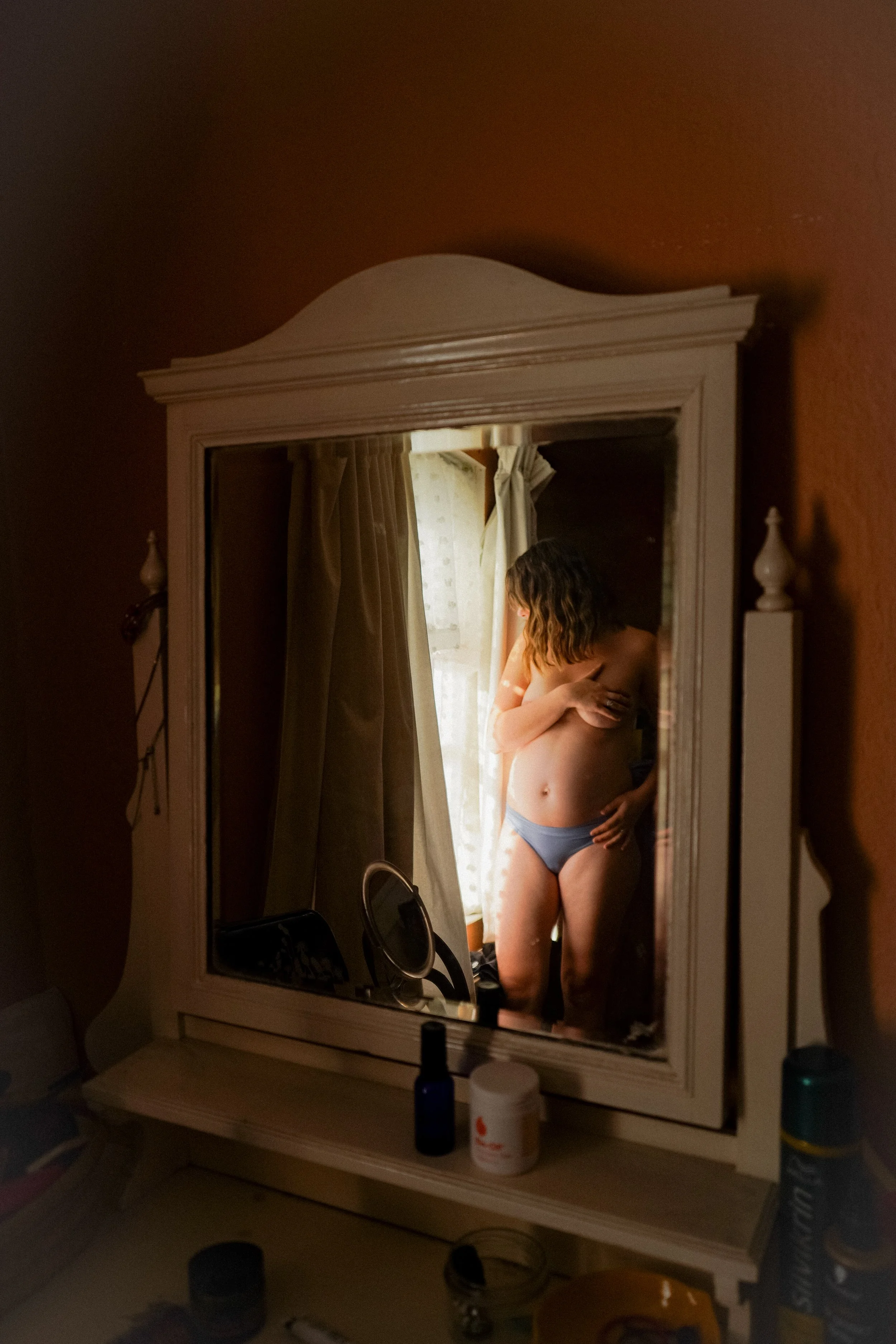 A woman with wavy hair standing in front of a mirror, partially undressed, with her arms crossed over her chest, in a softly lit bedroom.
