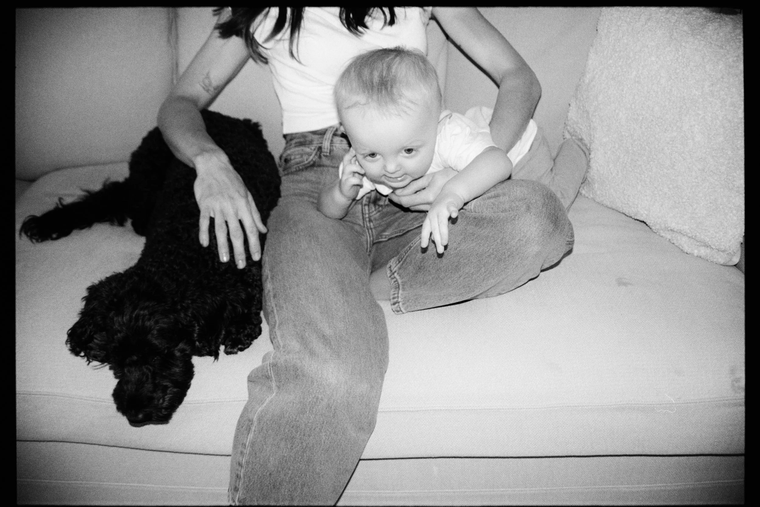 A person sitting on a couch with a small child on their lap and a large dog beside them. The person has long dark hair and is wearing a short-sleeved shirt and jeans. The child has short hair, is wearing a light shirt, and is playfully holding their 