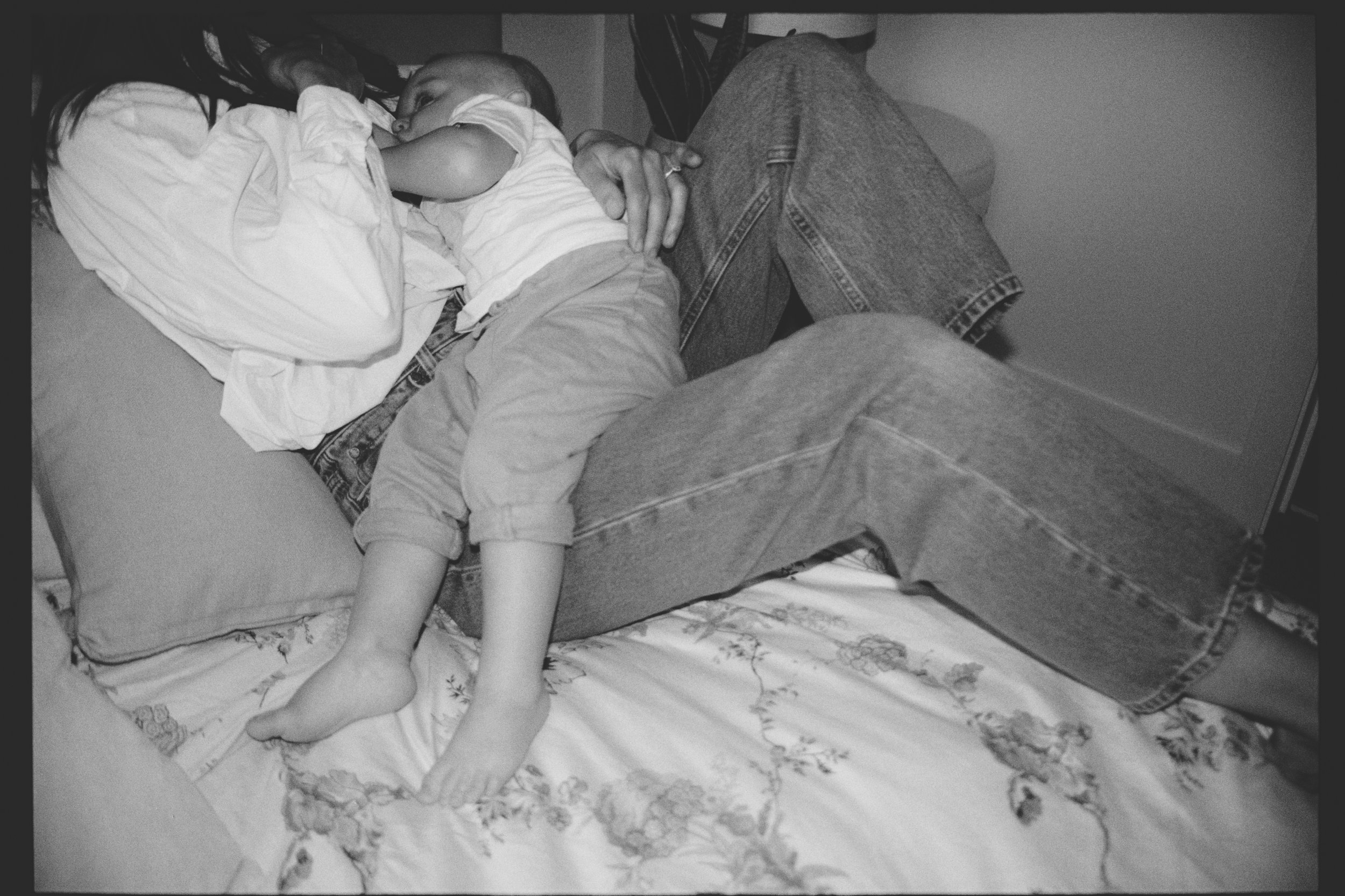 A grayscale photo of a young child lying on a bed, cuddling with an adult. The child wears a striped shirt and shorts. The adult is wearing jeans, and only part of their body is visible. The bed has a floral blanket, and there is a plain wall in the 
